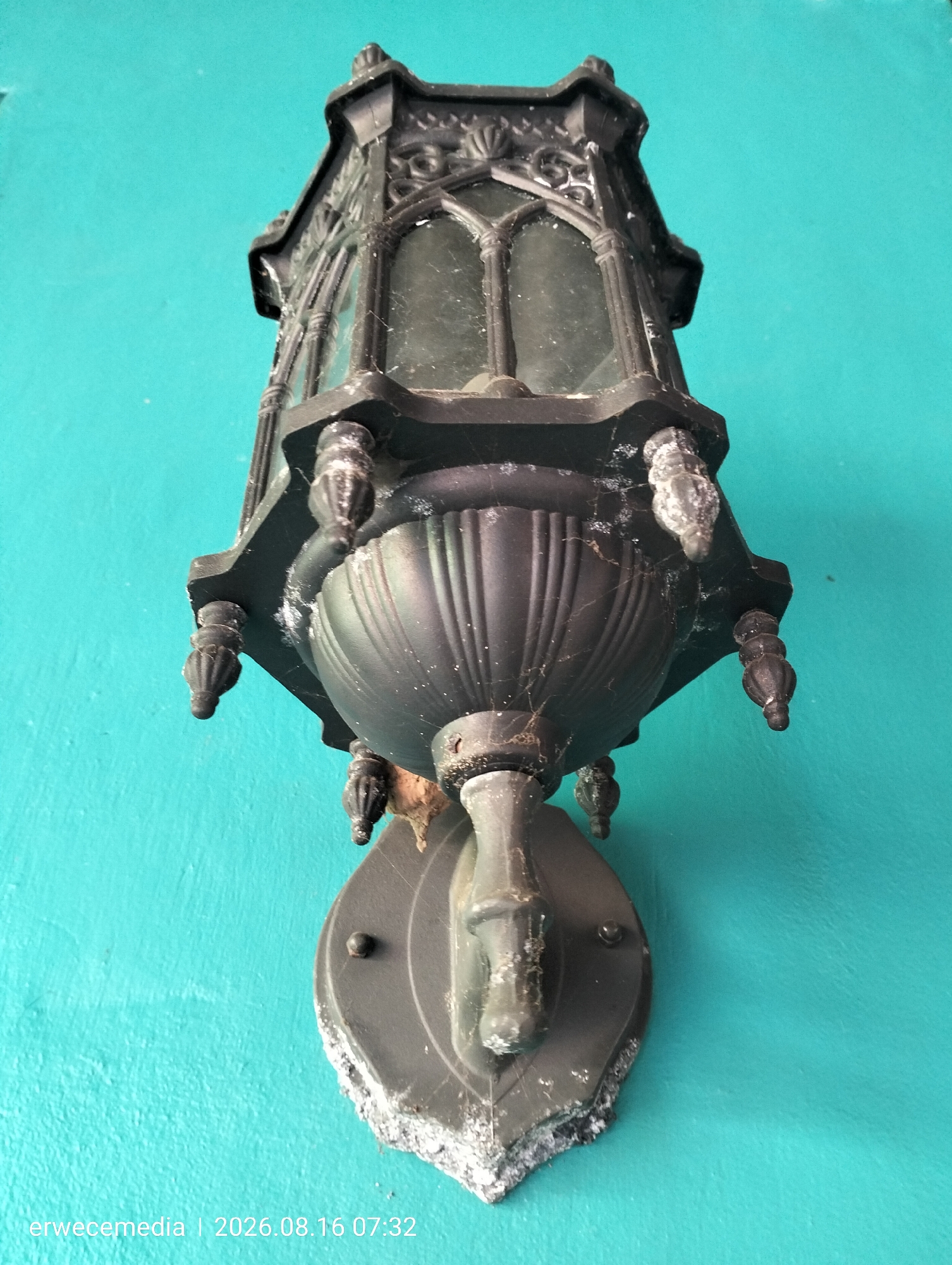 An ornate, black, cast metal wall lantern hangs on a teal-colored wall. The lantern has a Gothic architectural style with arched glass panes and decorative finials. The metal shows signs of age and wear, with some white debris and cobwebs visible. The bottom of the lantern is attached to a decorative metal mounting plate. A watermark with "erwecemedia | 2026.08.16 07:32" is present in the lower-left corner of the image. The location specified is the city of Imogiri, Indonesia.