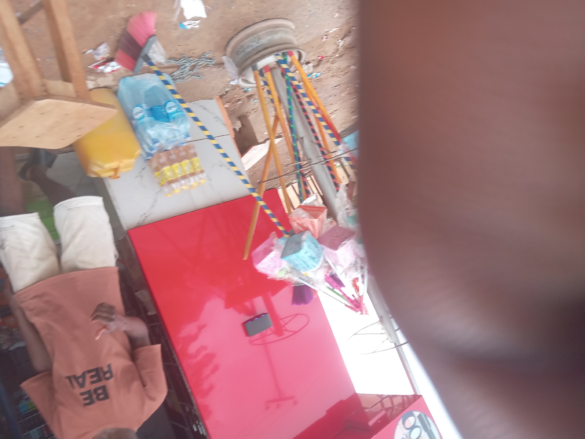 The image shows a scene in what appears to be a small business or workshop in Achara Ndiagu Omegio, Nigeria. A person wearing white shorts and an orange shirt with the words "BE REAL" is partially visible. On a red table, there is a mobile phone and assorted wrapped gift items. A container of drinking water, bottled juice, and other objects are placed on a white surface adjacent to the table. A wheel rim is supported by painted wooden poles in the background. The floor appears to be a mixture of tiles and dirt.