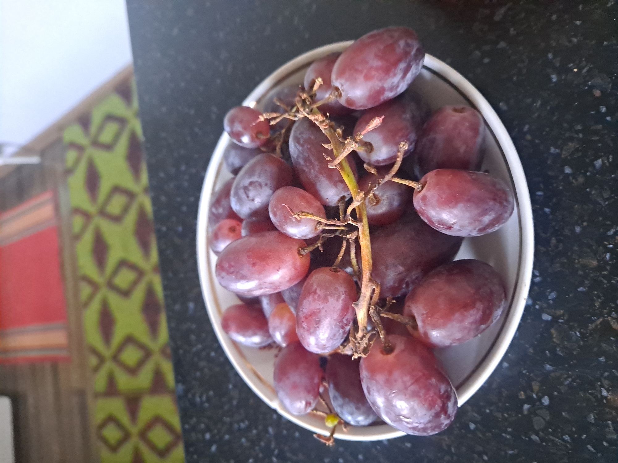 A bowl of red grapes sits on a dark countertop. The bowl is white with a thin gold rim. The grapes are plump and oval-shaped, with some appearing to have a dusty bloom on their surface. They are clustered together on a brown stem. The countertop is speckled with light-colored flecks. In the background, a patterned rug with green and brown diamond shapes is visible, along with a red rug. The image appears to be taken indoors, possibly in a kitchen or dining area.