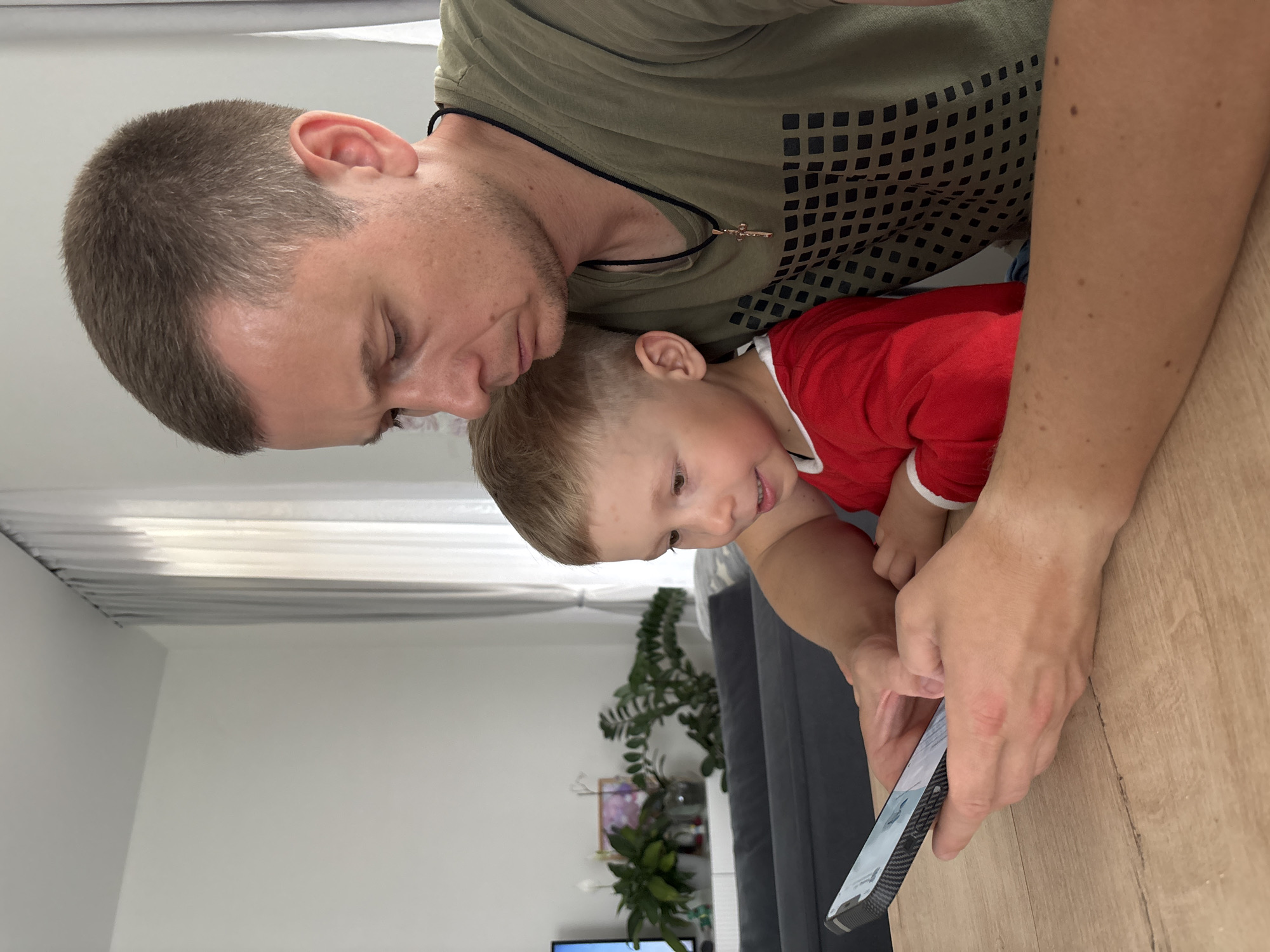 A man and a young child are looking at a mobile phone held in the man's hands. The man is wearing a green t-shirt with a dark geometric pattern and a black necklace with a cross pendant. The child is wearing a red shirt with white trim. They are positioned near a light-colored wooden table. In the background, there's a gray couch, a plant, a picture frame, and a television set. The room appears to be well-lit, with white walls and curtains. There is no information in the image that can confirm a specific location, such as Chaikovsky, Russia.