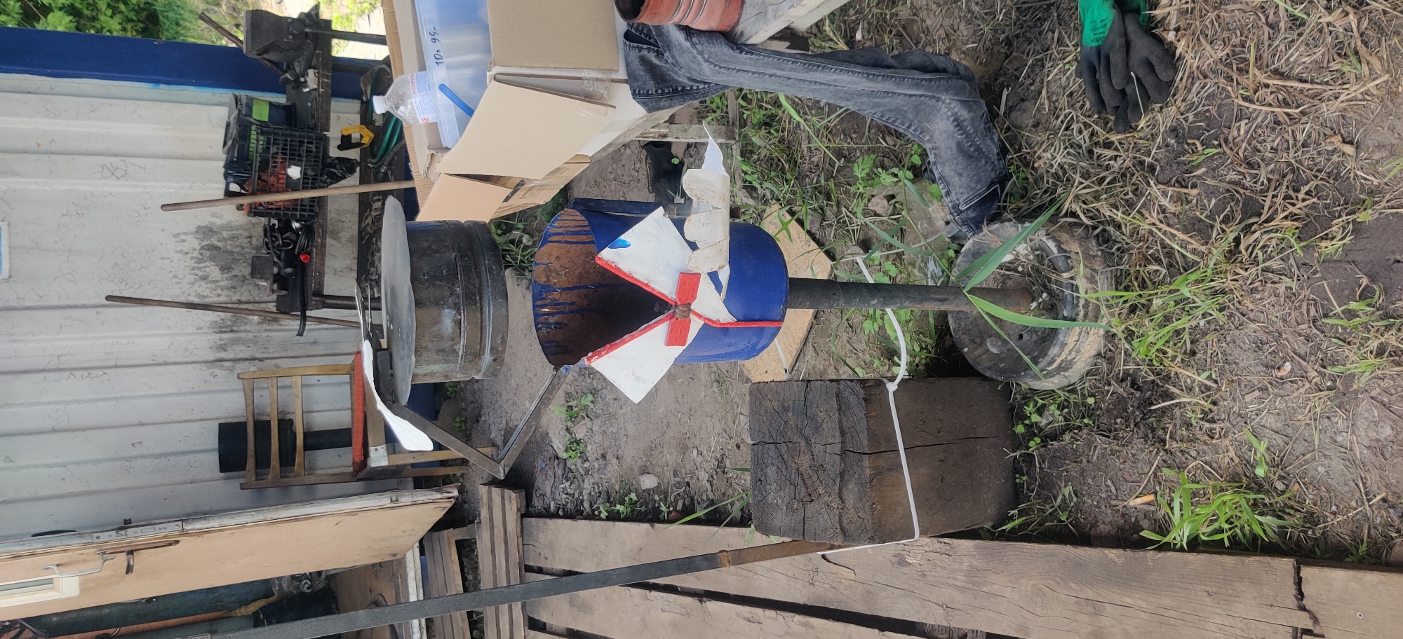 The image depicts an outdoor scene in Rizunenkove, Ukraine, likely during daytime under overcast or moderate lighting conditions. The primary subject is a homemade device resembling a wind turbine or a fan, constructed from a blue barrel with white and red blades attached to a central rotating shaft. This shaft is mounted on a stand that appears to incorporate a wheel, suggesting it might be mobile or intended for outdoor use.
Beside the wind turbine, a large, dark metal pot or cauldron is visible, positioned on a metal frame. In the background, there are various objects including cardboard boxes, a plastic bottle, a black plastic crate, and what seems to be a pair of green work gloves lying on the ground. The ground is covered with dirt and dry grass. The scene also includes wooden elements, such as planks and a thick log, which seem to be part of the surrounding environment or supporting structures.
A person's legs, clad in dark denim jeans and possibly wearing work boots, are visible on the right side of the image, suggesting human presence and activity. The overall setting appears to be a workshop, yard, or a rural area. There is no clear indication of a specific event taking place, but the arrangement of objects suggests ongoing work or a completed project. The image is oriented sideways, with the top of the frame pointing to the left. No visible text is discernable.