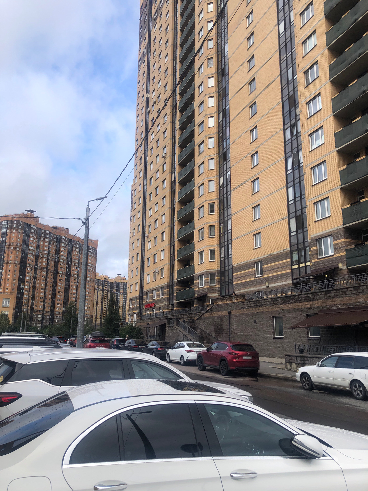 The image depicts a street scene in Pargolovo, Russia, dominated by tall, modern apartment buildings. A light beige and brown brick high-rise with balconies is prominently featured on the right, with dark green railing. To the left, another large apartment complex with a similar color scheme and numerous windows is visible. The sky is overcast with scattered clouds. Several cars are parked on the street and in a lot, including a white sedan in the foreground, a white station wagon, a red SUV, and other vehicles in the background. A street lamp with wires runs across the image. The buildings appear to be residential complexes.