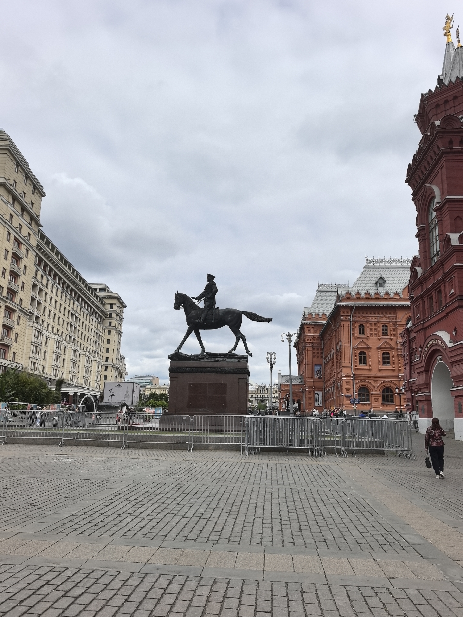An outdoor, wide-angle shot captures a historical city square in Moscow, Russia, under an overcast sky. In the foreground, the ground is paved with grey cobblestones, intersected by sections of lighter-colored paving. A series of silver metal crowd control barriers extends across the midground, separating the viewer's space from the central monument and surrounding buildings.
The focal point is a large bronze equestrian statue of a man in uniform, likely Marshal Georgy Zhukov, positioned on a dark, rectangular pedestal in the center. The rider faces right, mounted on a horse with its front left leg raised.
To the left of the statue stands a grand, multi-story building with a light-colored facade, numerous windows, and classical architectural details, characteristic of the Hotel National. To the right, a distinctive red brick building with ornate white trim, grey roofs, and golden finials dominates the scene. This is the State Historical Museum, with the words "ИСТОРИЧЕСКИЙ МУЗЕЙ" (Historical Museum) partially visible above an arched entrance on its right side.
Few people are visible; one woman walks away from the viewer on the far right, and a few indistinct figures can be seen behind the barriers on the left. The cloudy sky suggests a calm, possibly cool, daytime setting. The presence of the barriers implies that the area may be prepared for an event or has recently hosted one. The overall atmosphere is quiet and monumental, showcasing iconic Russian architecture and a significant historical landmark in Moscow.