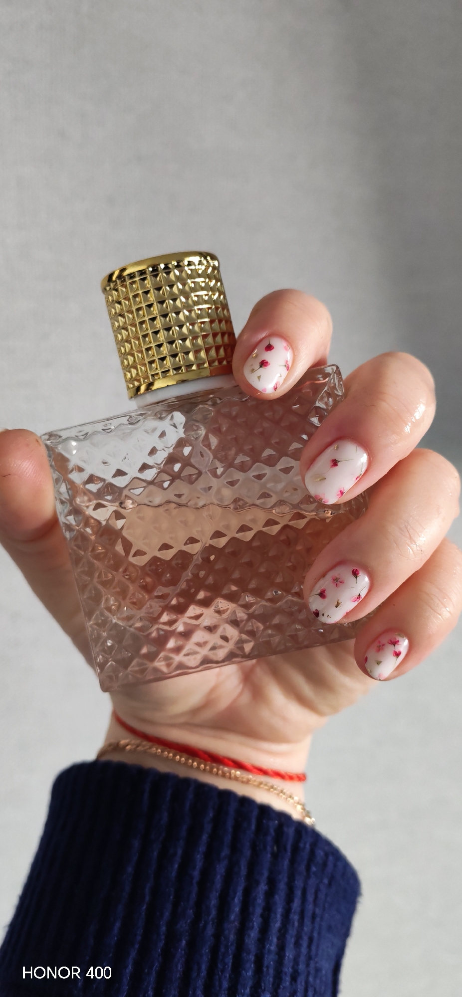 A hand holds a clear, textured glass perfume bottle with a gold, latticed cap. The nails are painted white with small, scattered floral designs. The hand wears a blue sweater and two bracelets, one gold and one red. The background is a plain gray wall. The text "HONOR 400" is visible in the lower left corner.