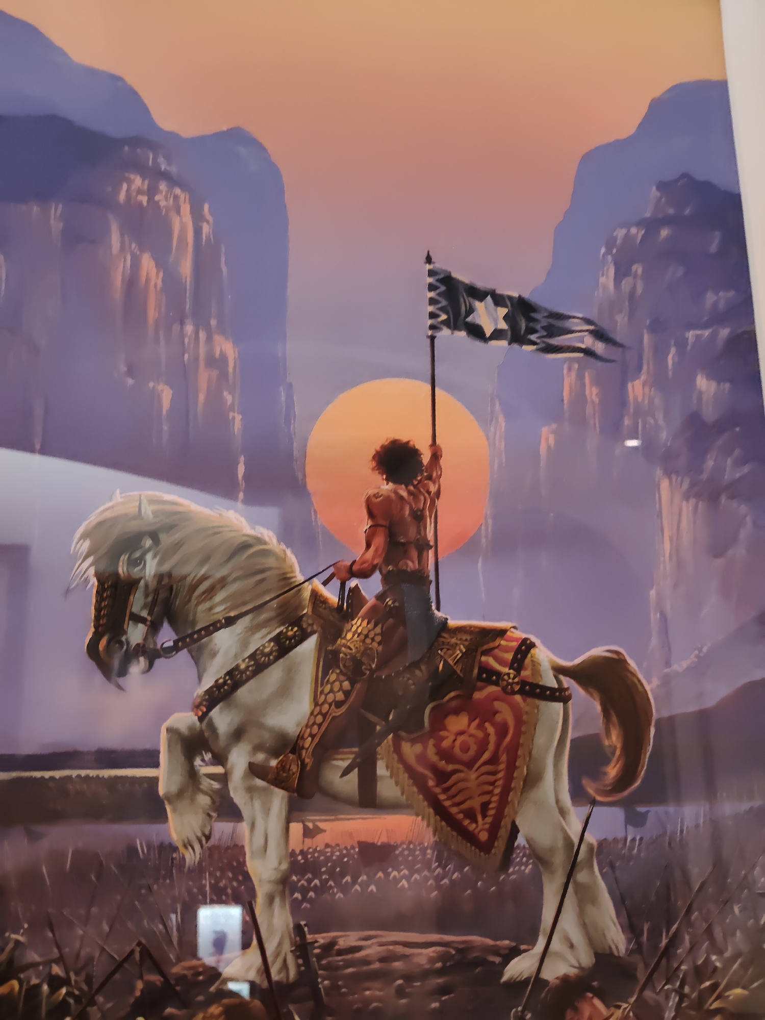 This is a photograph of a painting depicting a warrior on horseback in a dramatic, mountainous landscape at sunset. The warrior, seen from behind, wears minimal armor and holds a black and white flag with a star design aloft. The horse is white with elaborate decorative tack. In the foreground, a vast army of silhouetted soldiers stands at attention. The background features towering, rugged mountains bathed in the warm hues of a setting sun. The style of the painting suggests a fantasy or historical epic theme. The location is not specified within the image itself; it is a photograph of a painting, and the painting's content does not provide geographical context.