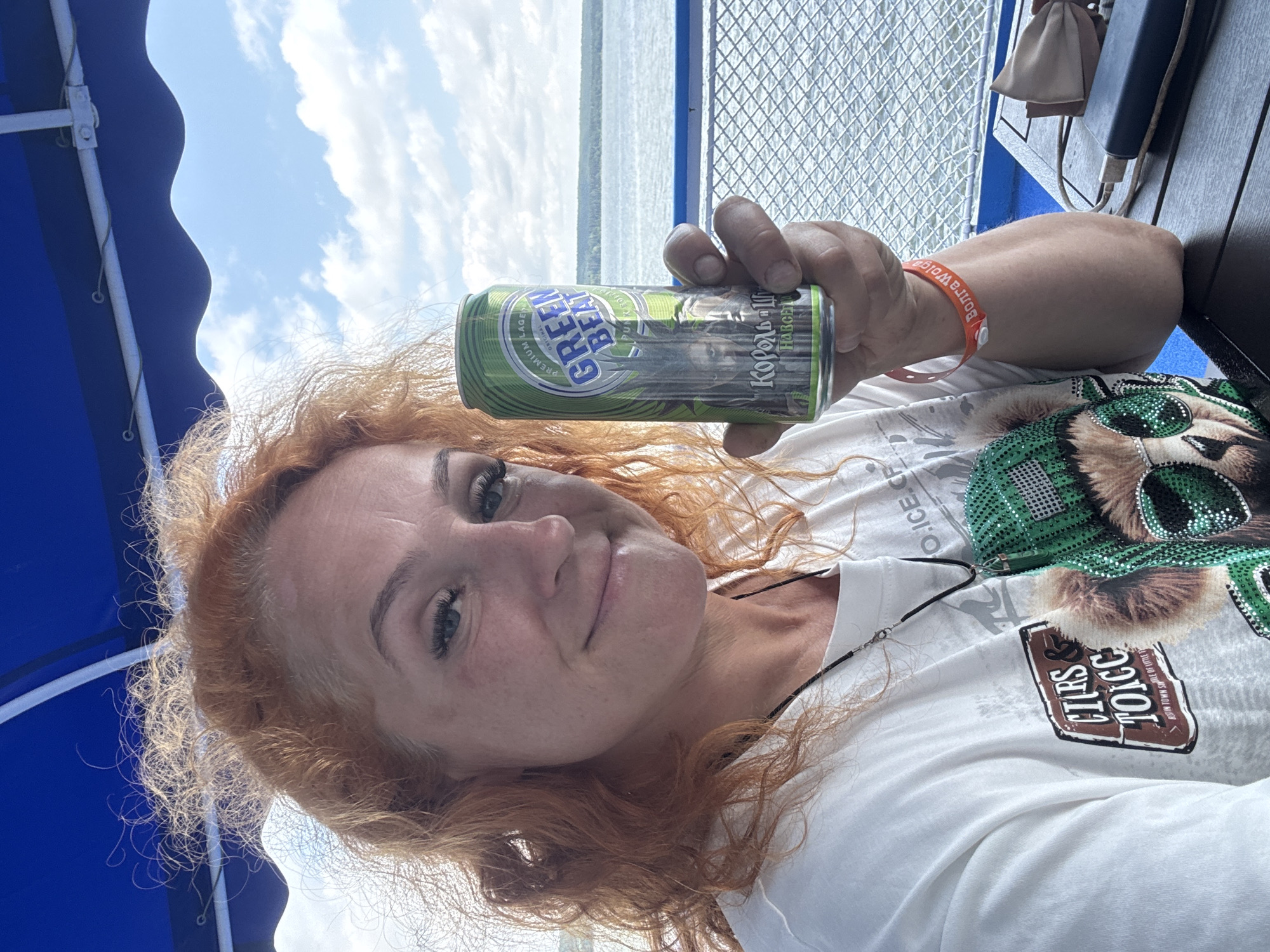 A woman with curly red hair is holding a can of Green Beat beer in her right hand, looking at the camera. She is wearing a white t-shirt with a graphic print. She has on a black necklace. In the background, there is a body of water with a blue railing and a metal fence, suggesting she is on a boat. The sky is visible with scattered clouds. The location is identified as Shelanga, Russia.