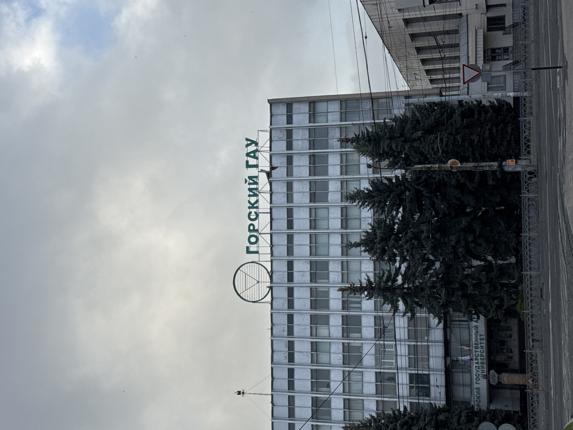 The image shows a building in Akhieli, Georgia. The building has a sign on the top that reads "ГОРСКИЙ ГАУ" in green letters. Below that, there is a round symbol. There are trees in front of the building. A road and a sidewalk are on the right side of the image. The sky is overcast. A sign with a red border stands at the street corner.