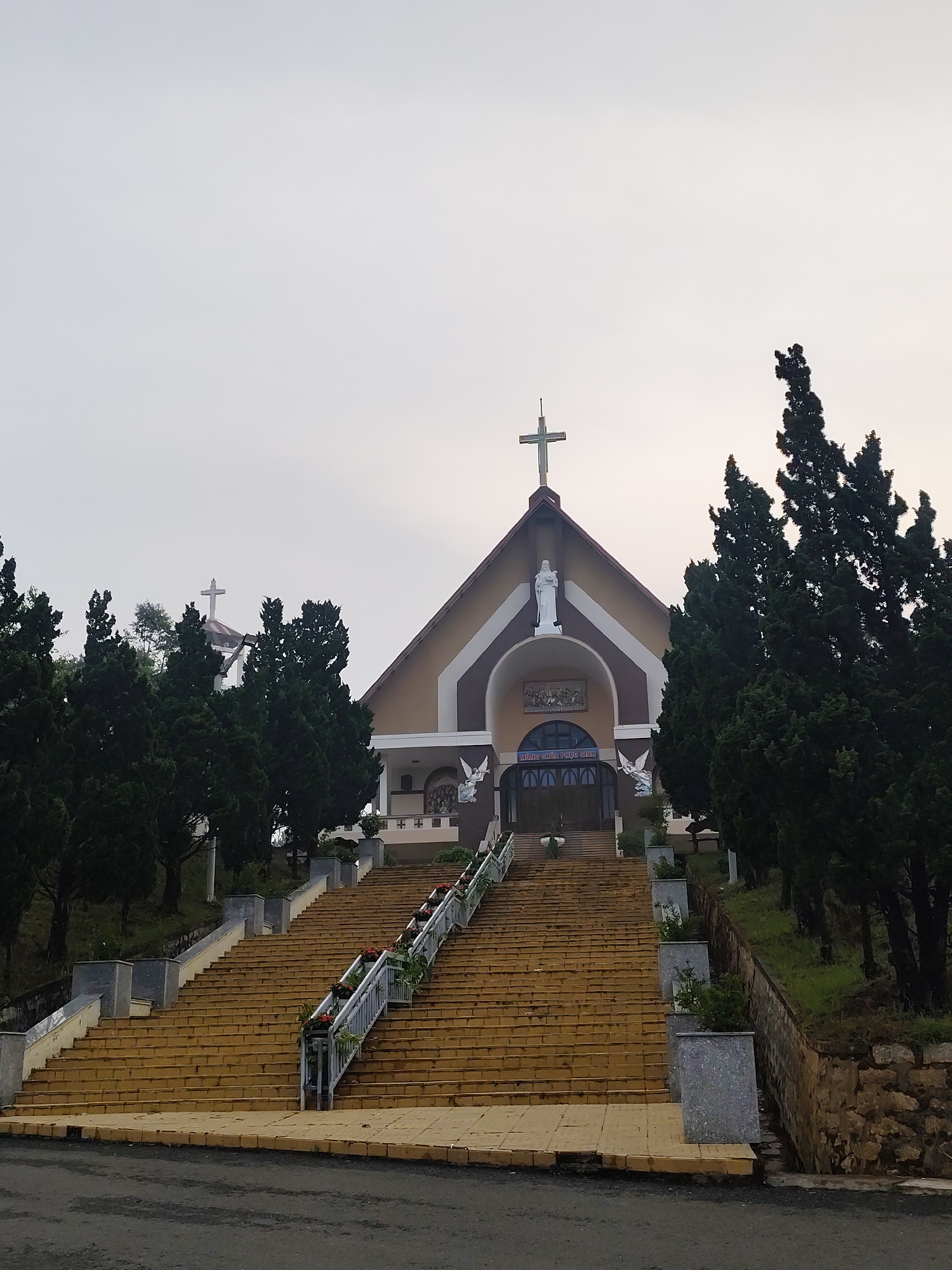 The image displays a Catholic church prominently situated atop a hill, accessed by a long and wide flight of yellow-brown tiled stairs. A large cross crowns the church's main roof, and another cross is visible in the distance behind trees to the left. Above the central entrance of the church, a white statue of the Virgin Mary stands, with two white angel statues flanking the arched doorway below.
Above the entrance, a sign displays the Vietnamese text "MỪNG CHÚA PHỤC SINH," which translates to "HAPPY EASTER" or "CHRIST IS RISEN." The church building itself features a gabled roof and is primarily beige and brown in color.
Dark green, tall coniferous trees, possibly cypress, densely frame both sides of the ascending stairs and surround the church, lending a lush and secluded feel to the setting. The long staircase is equipped with metal railings and adorned with planters containing red flowers and greenery. Granite-like planters and stone retaining walls line the sides of the stairway and the surrounding landscape.
In the foreground, a section of an asphalt road is visible. The sky is overcast and light grey, suggesting either a cloudy day or early morning/late afternoon light. The overall scene is static and tranquil, depicting the architecture and natural environment. This location is identified as being in Lang Biang, Da Lat, Vietnam.