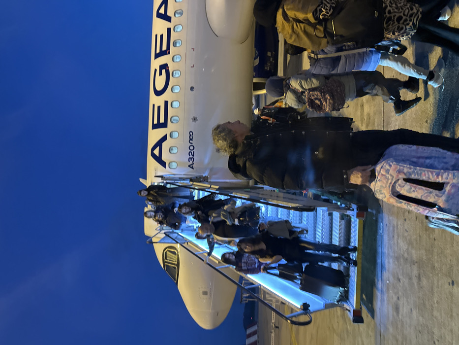 People are disembarking an Aegean A320neo aircraft via stairs. Several passengers, including some with luggage, are visible on the steps. The plane's fuselage, with "AEGEAN" printed in blue, is partially visible in the background, along with a blue sky. The image is taken from a close perspective, capturing the moment of arrival or departure.