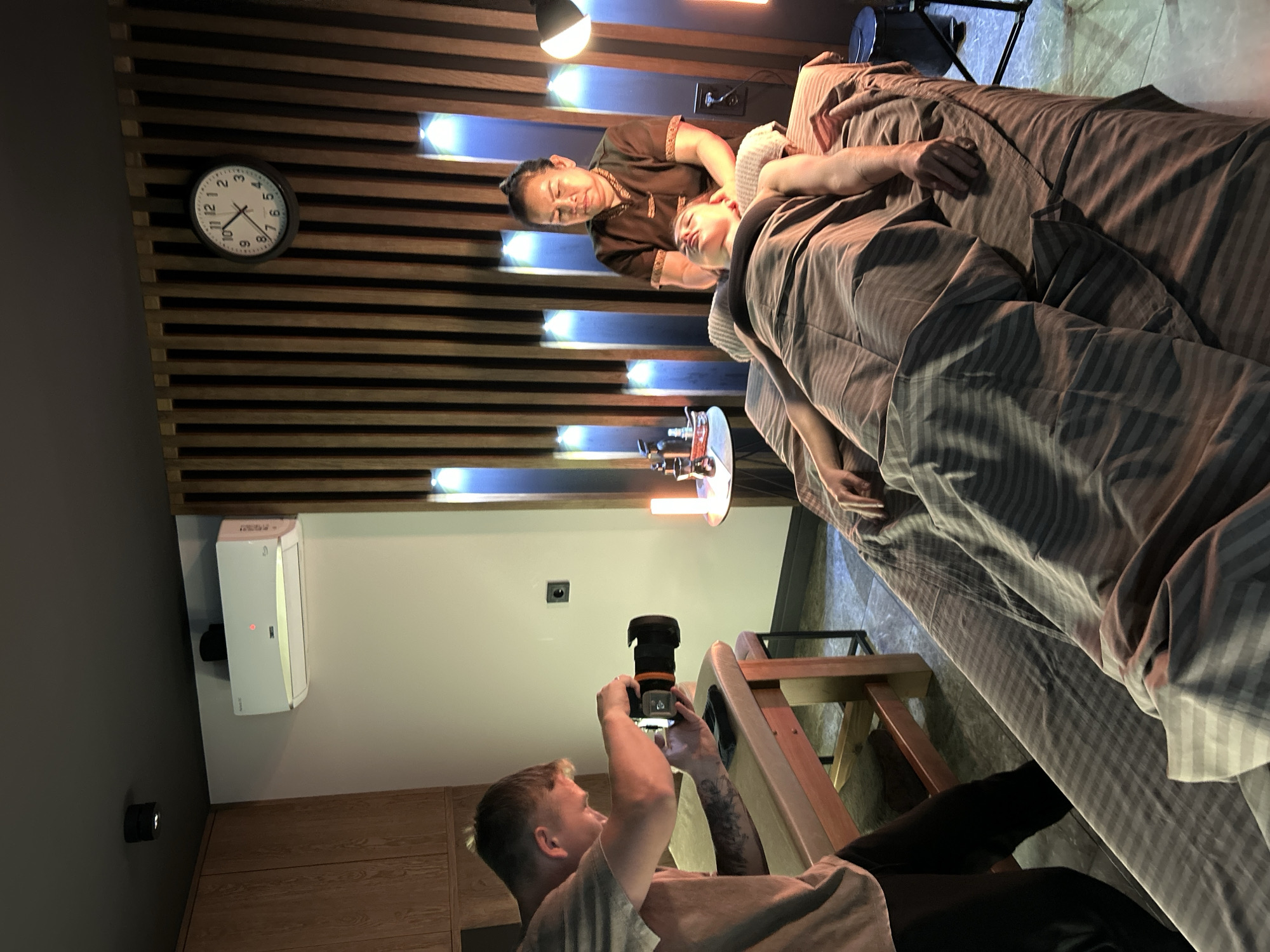 A man is taking a photo of a woman receiving a head massage. The massage is being given by another woman. The scene is indoors. There is a clock on the wall above the massage area showing 10:08. There is an air conditioning unit on the wall. Location: Votkinsk, Russia.