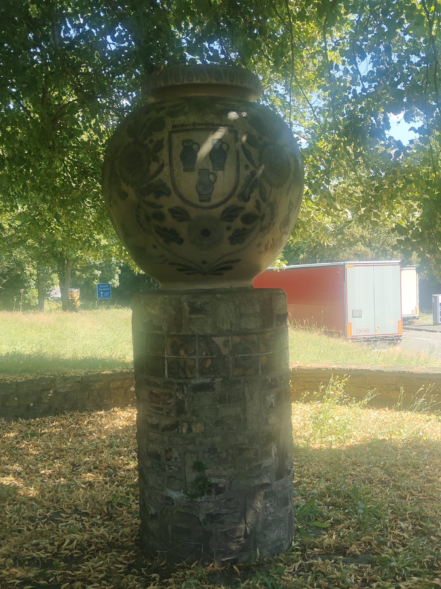 The image shows a large decorative pot, likely made of stoneware, displayed on a brick pedestal in an outdoor setting. The pot is round and tapers towards the top, featuring intricate blue and white decorative patterns and a crest with three pitchers. The pedestal is constructed from weathered bricks and appears to be cylindrical. The scene is set outdoors with lush green trees providing shade over the monument. In the background, there is a grassy area with a low stone wall, a road with several parked semi-trailers, and a blue directional signpost indicating "Dreieck Buchenau". The ground around the pedestal is covered with fallen leaves and some sparse vegetation. The lighting suggests it is a sunny day with dappled sunlight filtering through the leaves. The overall impression is of an art installation or historical marker located in a public space, possibly a park or roadside in Höhr-Grenzhausen, Germany, given the directional sign.
