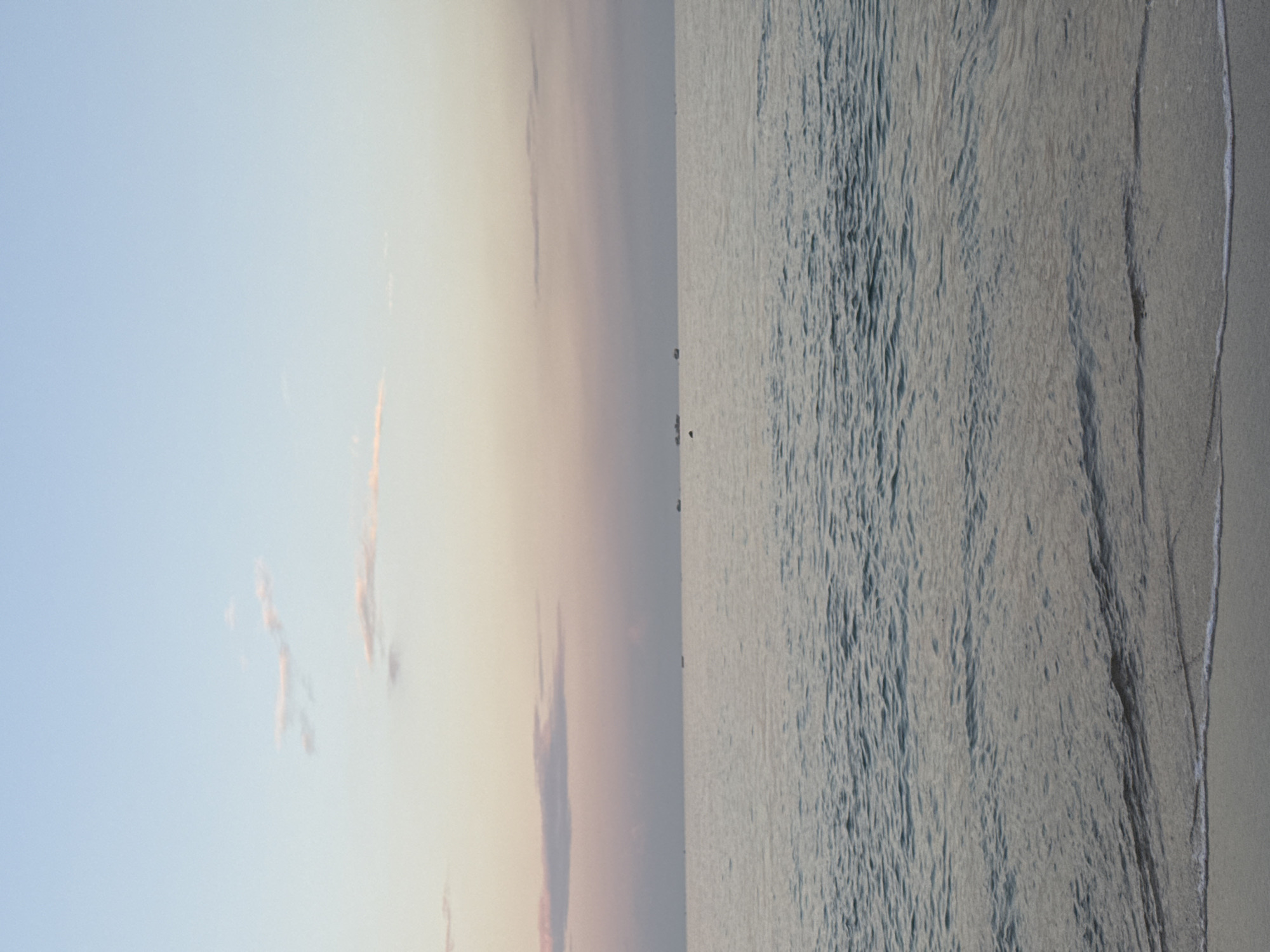 The image depicts a beach scene, likely in Kuta, Indonesia. The left side features a sky with a gradient from light blue to a soft, pale orange near the horizon, containing thin, wispy clouds tinged with pink. The right side of the image shows the ocean surface with subtle wave patterns, transitioning to a sandy beach on the right edge.