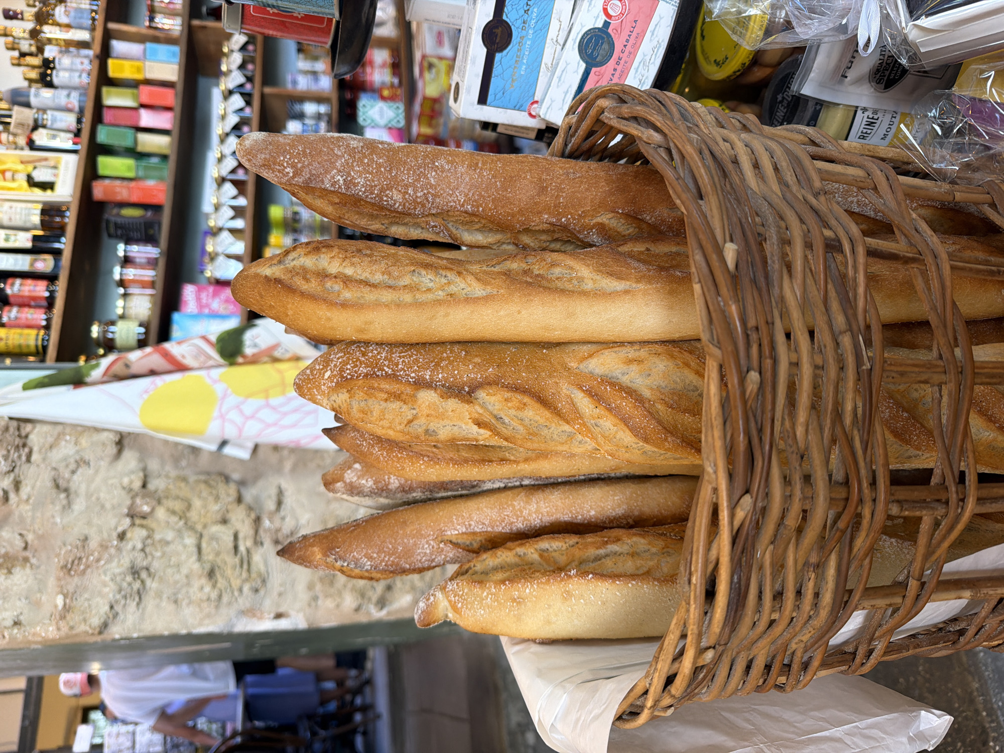 In a Tel-Aviv bakery, a woven basket overflows with golden-brown baguettes dusted with flour. The baguettes are long and crusty, with visible scoring marks on their surfaces. Behind the basket, shelves are stocked with an assortment of jars, bottles, and packaged goods, including what appear to be preserves, sauces, and other food items. A white and yellow patterned cloth hangs on the left side of the frame. In the background, a person wearing a white t-shirt and a pink cap is visible, along with a blurred background of what seems to be a marketplace or shop interior.