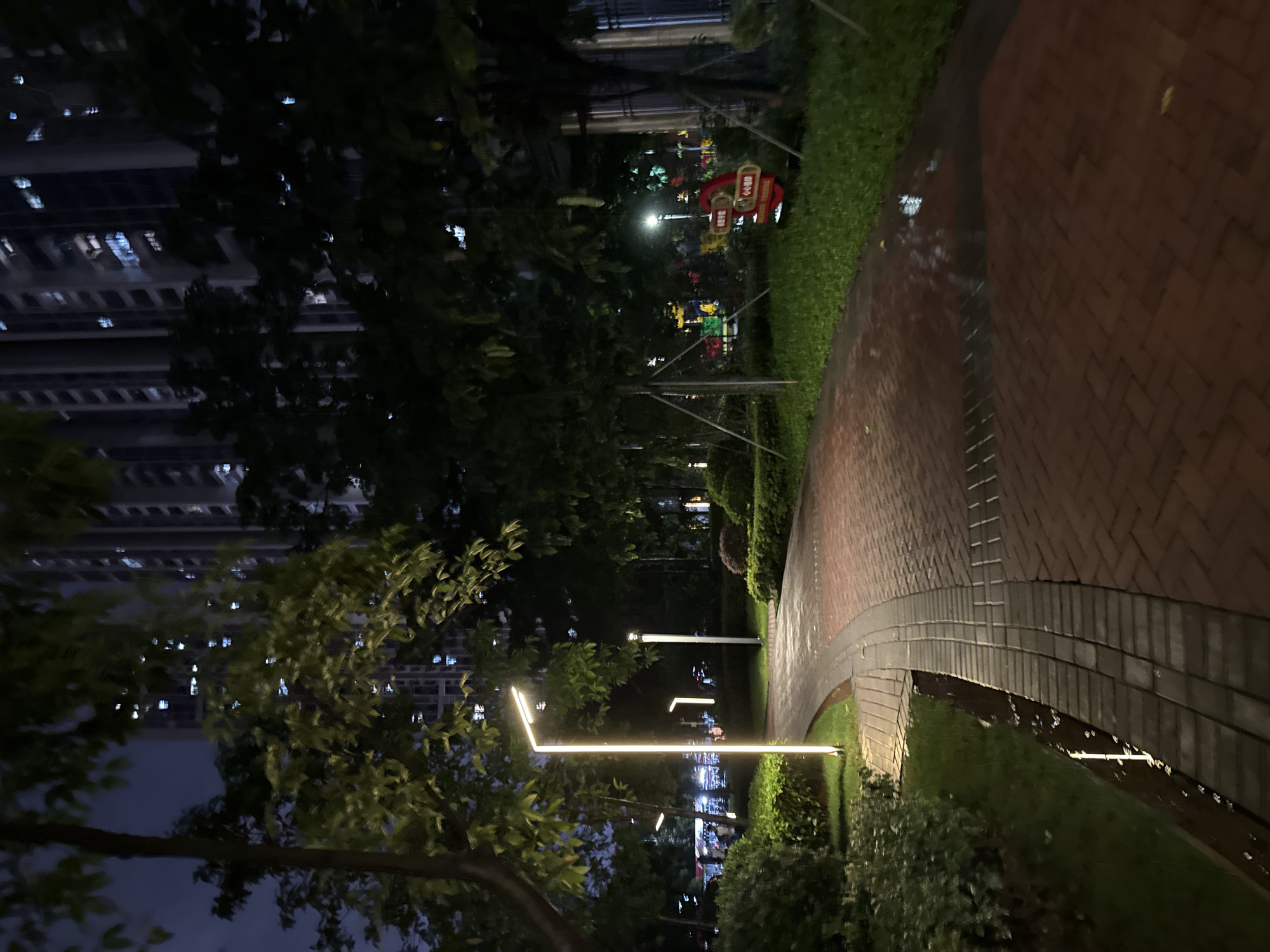 In Haizhu District, China, at night, a park pathway made of red bricks curves through a green space. The path is wet, reflecting the ambient light. Modern lampposts with warm, yellow light illuminate the walkway. Lush green bushes and trees line the path, with some trees having visible support structures. In the background, a tall, modern building with many illuminated windows stands against the dark sky. The overall scene depicts a well-maintained urban park at night, likely after a rain shower. There are no people or animals clearly visible in the frame.