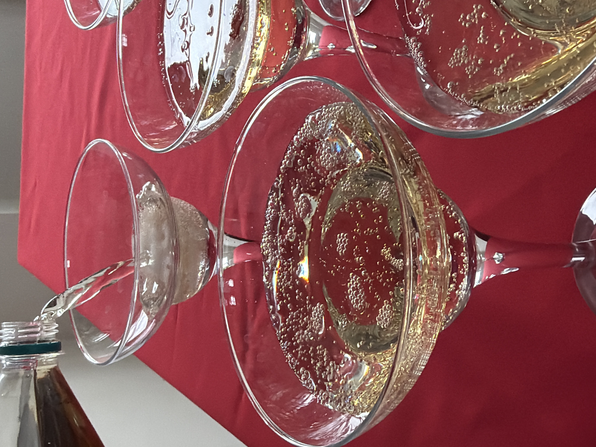 An indoor scene in Chaikovsky, Russia, depicts a close-up view of a beverage service setup, likely a champagne tower, positioned on a red tablecloth. Multiple clear glass coupe-style champagne glasses are arranged in a tiered formation. A clear glass bottle, partially visible on the upper left, is actively pouring a stream of clear, effervescent liquid into the topmost visible glass. This liquid cascades into lower glasses, which are already partially filled with a pale golden, bubbly beverage. The liquid exhibits numerous small bubbles, particularly concentrated along the glass rims and within the liquid itself, creating refractive light patterns. The background consists predominantly of the red tablecloth, with a portion of a plain, light-colored wall or surface visible on the far left. No individuals are present in the frame.