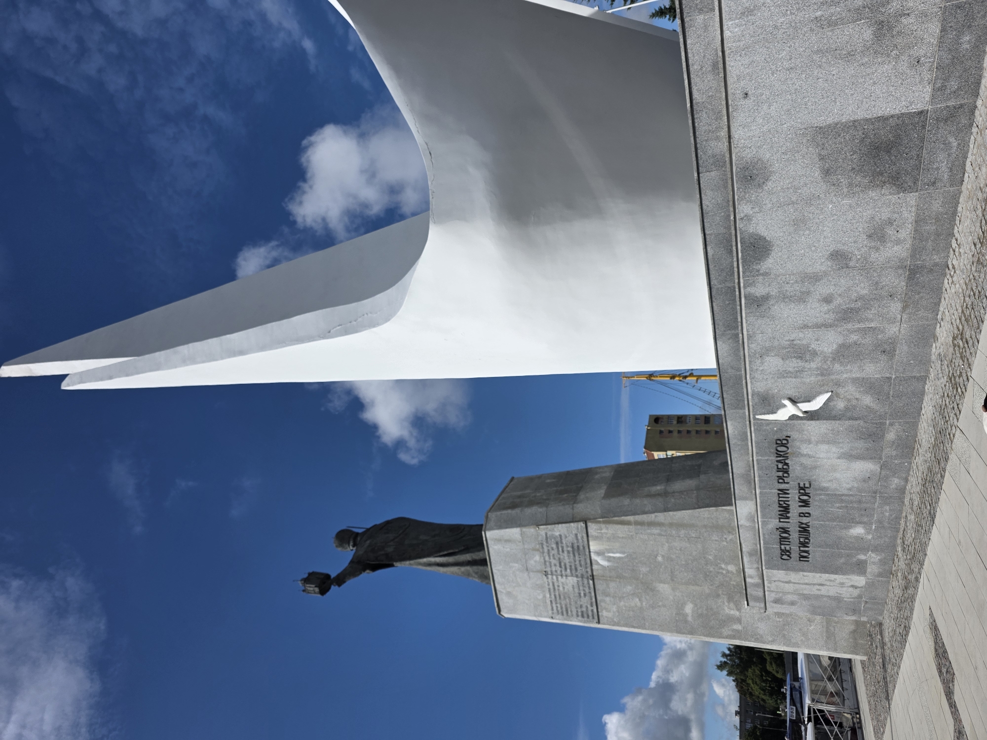 A statue of a fisherman holding a lantern aloft stands on a grey granite pedestal. The statue is positioned to the left of a large, modern, white sculpture that curves upwards against a bright blue sky with scattered white clouds. The base of the monument features an inscription in Russian, which translates to "In memory of fishermen who perished at sea." A small white bird sculpture is mounted on the granite base to the right of the inscription. In the background, a tall residential building and part of a ship or boat can be seen. The ground in front of the monument is paved with grey and light-colored tiles and cobblestones.