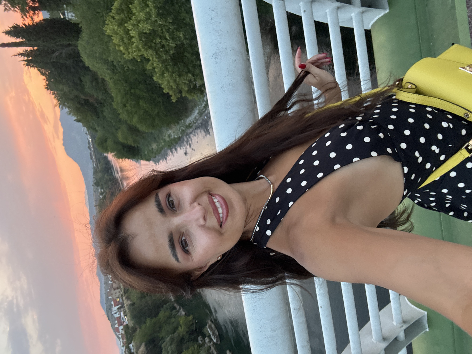 An outdoor, eye-level selfie shows a woman on a bridge with a river and trees in the background. The woman has long, dark hair and is wearing a black and white polka dot dress and a yellow purse. She is smiling and holding a strand of her hair. The sky is a warm pink and orange, suggesting sunset. The bridge has white railings.