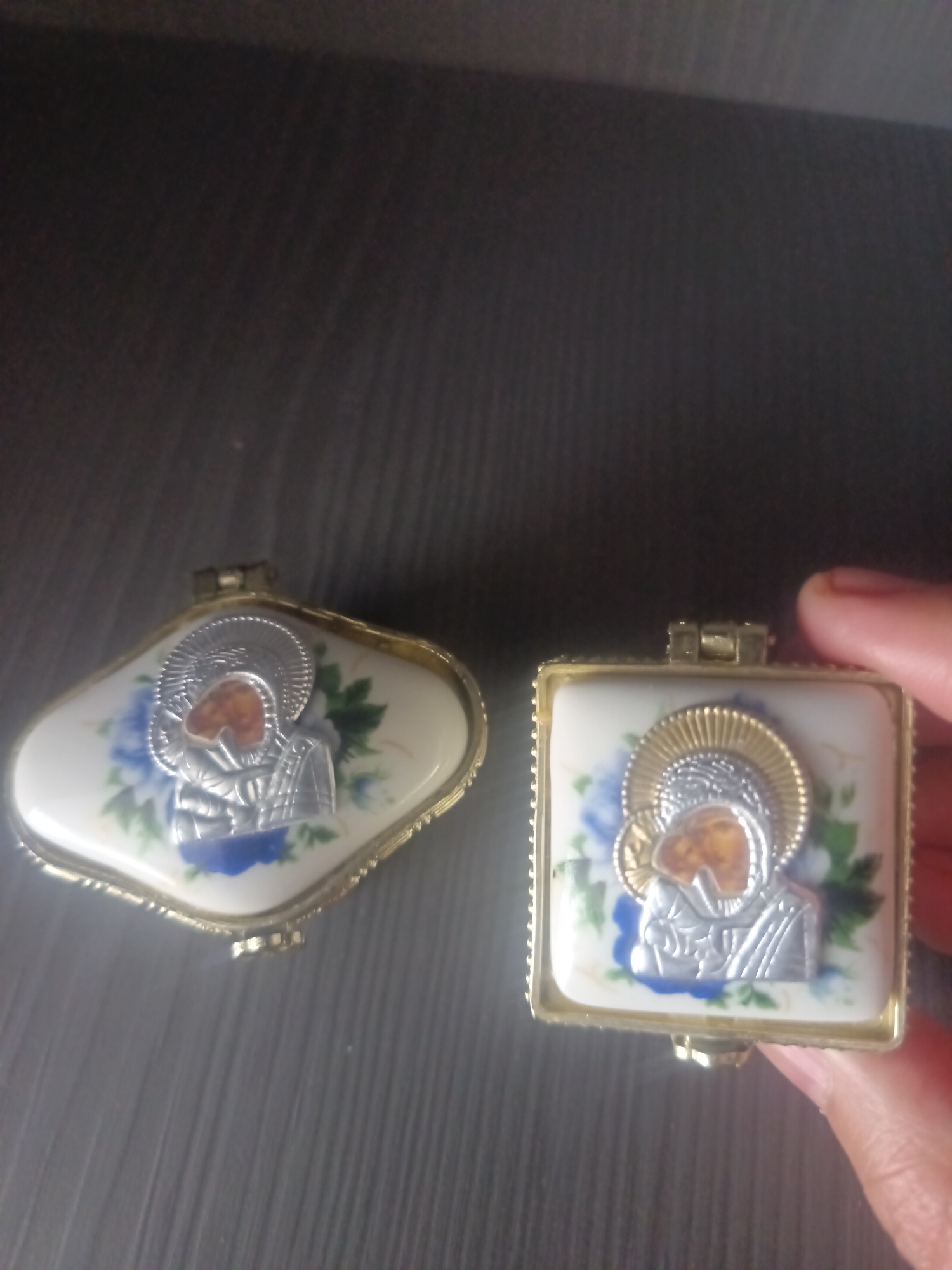 Two small, ornate lidded boxes are displayed on a dark, textured surface. The box on the left is irregular in shape, resembling a teardrop or a plump oval, and is edged with gold-toned metal. Its surface is white porcelain adorned with a floral pattern of blue and green flowers and leaves. Centered on the lid is a raised silver and gold religious icon depicting a profile of a figure with a halo, featuring an amber-colored insert in the face area. The box on the right is square with rounded corners, also with a gold-toned metal edge. It shares the same white porcelain base with blue and green floral decorations. A similar silver and gold religious icon with an amber facial insert is centered on its lid. A person's hand, with visible fingernails, is partially visible on the right side of the image, gently holding the edge of the square box. The background is dark and out of focus, suggesting an indoor environment.