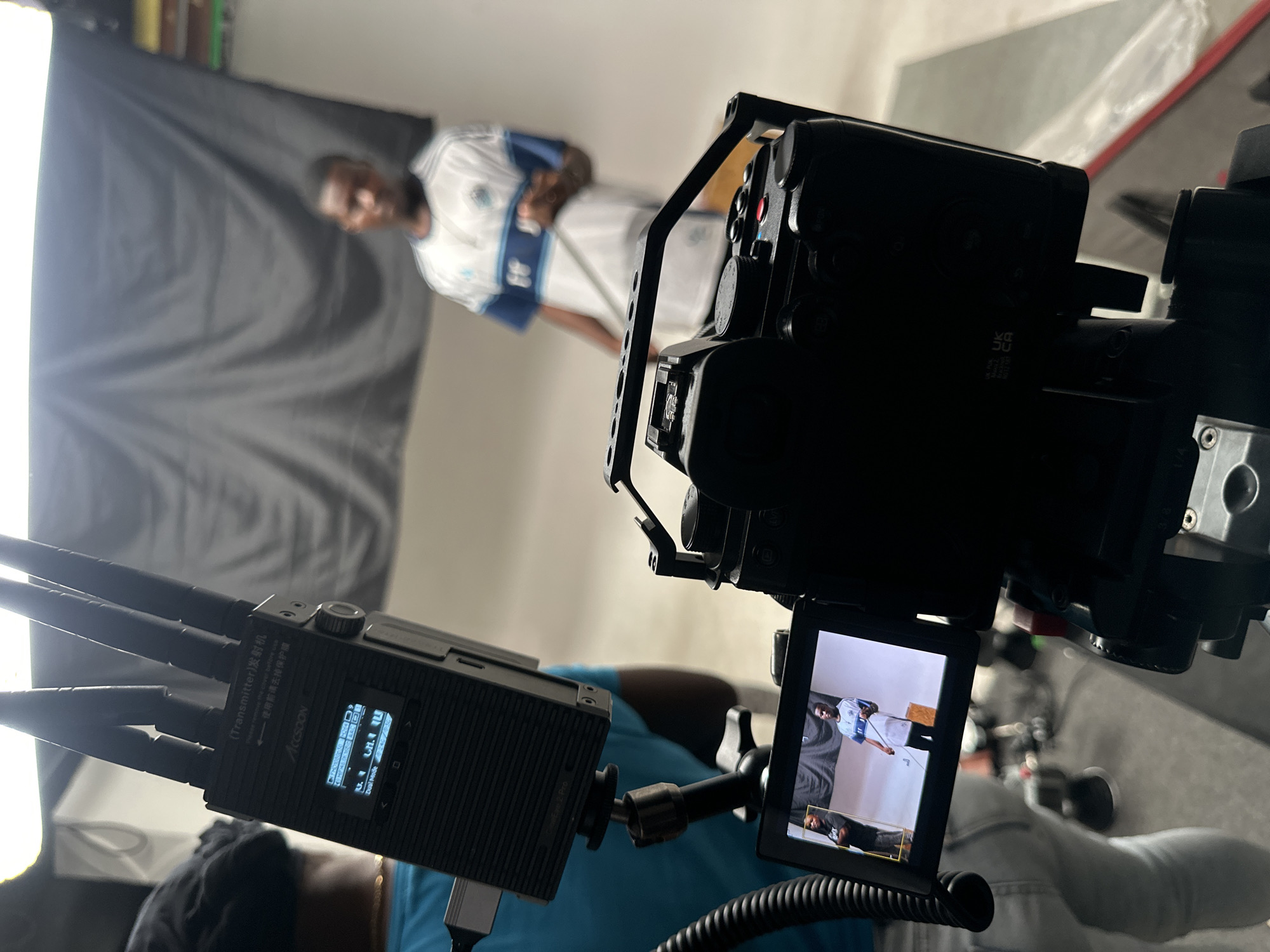 The image depicts an indoor video or photo shoot setup in Ikeja, Nigeria, captured from behind the camera equipment. In the immediate foreground, to the left, is a black Accsoon wireless video transmitter featuring three antennas. Its screen displays technical details such as "ACCSOON," "FHD1080P50," "Dual Mode," and "CH.1." Chinese text reading "(Transmitter) 发射机" and "使用前请去掉保护膜" (Please remove protective film before use) is visible on its casing.
To the right, a professional video camera, possibly housed in a cage, is the main focus. Its articulating monitor displays a live feed of the subject being filmed: a dark-skinned person, slightly out of focus, wearing a white and light blue patterned shirt, standing against a large dark grey or black fabric backdrop. A smaller frame on the monitor shows a different shot of a person lying down. Markings like "4K" and "DC" are partially visible on the camera body.
The scene suggests a controlled studio environment, illuminated by artificial lighting, with a softbox or similar light source visible to the far left, providing illumination for the subject. The overall setup indicates a professional video production or photography session in progress.