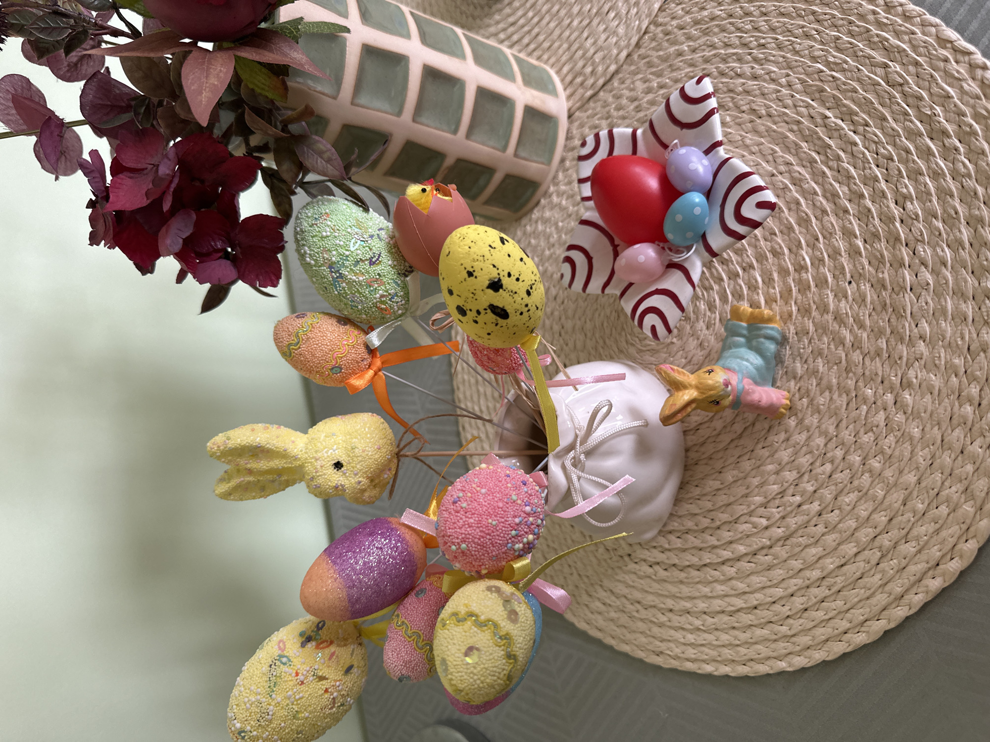 The image depicts a still life arrangement, likely indoors, featuring Easter-themed decorations. A bouquet of artificial flowers with deep red and green foliage is positioned on the left side of the frame, near a pale green decorative vase with a grid pattern. Adjacent to the flowers is a white vase containing several decorative Easter eggs and a small yellow rabbit. The eggs are varied in color and texture, some speckled, glittered, or wrapped with ribbon. To the right, a woven placemat provides a backdrop for a decoration of a red egg surrounded by smaller pastel eggs. A small bunny figure with pink and blue clothing lies on the placemat, and the scene is situated on a grey surface. The environment suggests a festive, celebratory atmosphere. There is no indication of Montana, Bulgaria within the image.