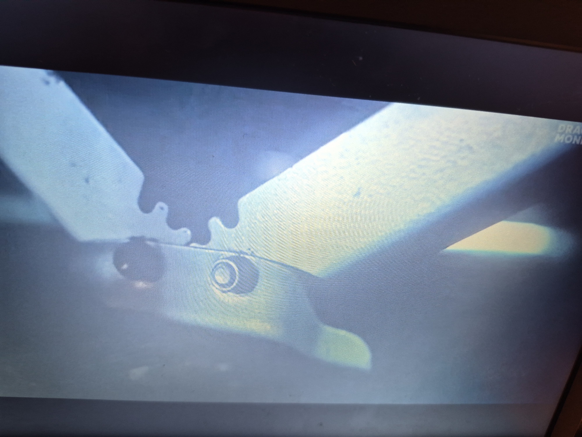 The image shows a close-up, blurry view of mechanical components, possibly from a machine or a device. There are metallic parts with visible rivets and a geared edge. The lighting is uneven, casting shadows and highlights. In the upper right corner of the image, a partial text overlay is visible, reading "DRAG" and "MONI", which might indicate a brand name or a date/time stamp. The background is out of focus and appears to be dark. There is no information in the image that allows for the identification of a specific location in Sandwell, United Kingdom.
