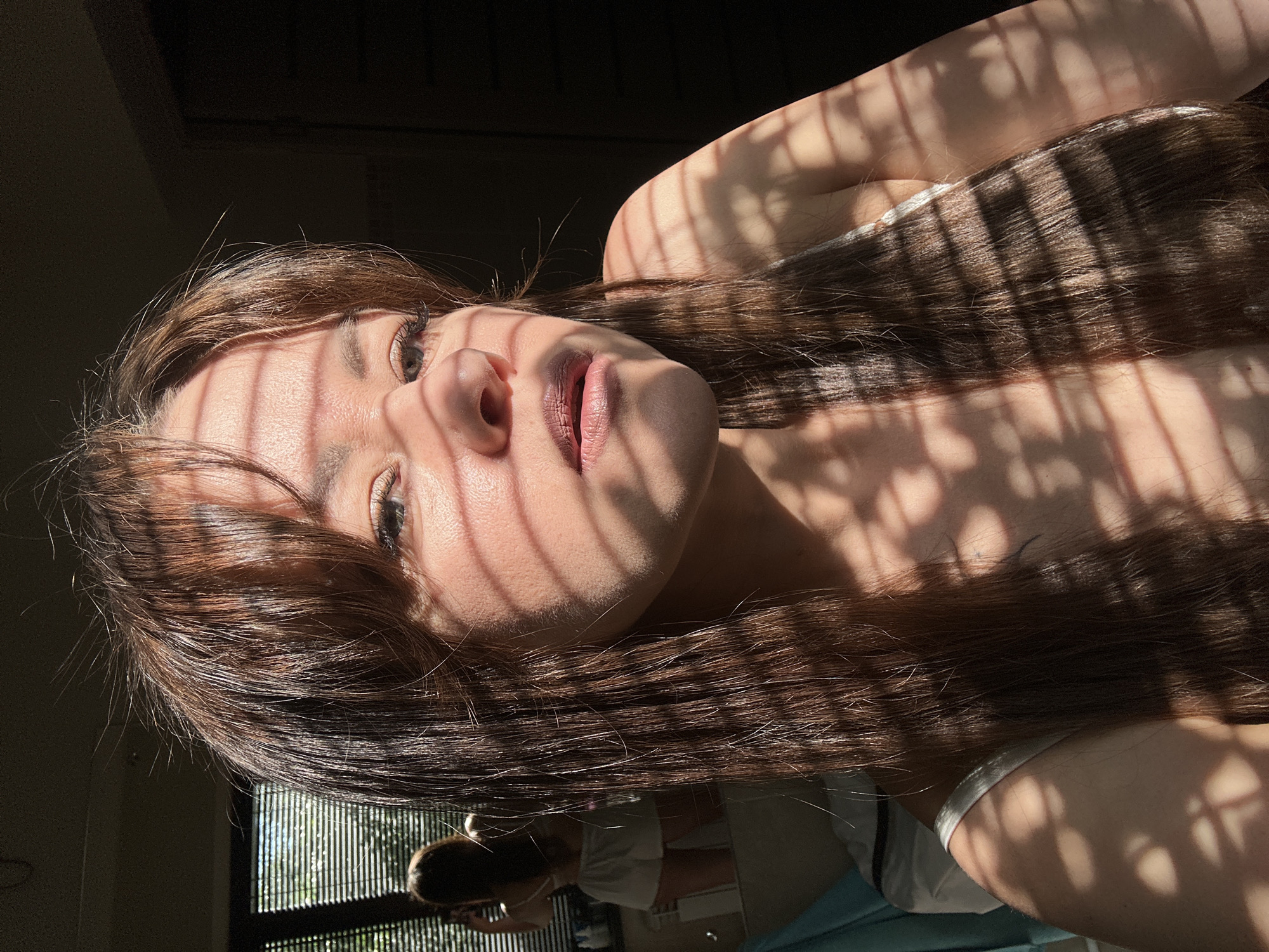 A young woman with long, brown hair is shown in a close-up, head-and-shoulders shot. She is looking towards the camera, with sunlight casting strong horizontal shadows across her face and body from what appears to be blinds. Her makeup is visible, with mascara and lipstick applied. She is wearing a light-colored top, possibly a camisole. In the background, a blurred reflection in a mirror shows the woman from behind, wearing white shorts and a similar light-colored top, suggesting she is in a bedroom or dressing area. The lighting suggests it is daytime, possibly morning or afternoon, with sunlight streaming in from a window. The environment appears to be indoors, with hints of furniture and possibly a window with blinds. There is no indication of the specific location within the city of Chaikovsky, Russia.