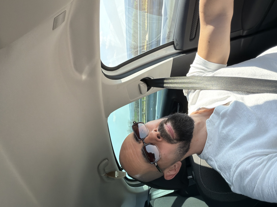 An adult male with a bald head and a trimmed dark beard is depicted seated in the driver's position of a vehicle. He wears dark-framed sunglasses and a white, short-sleeved, collared shirt. A dark-colored seatbelt is engaged across his chest and lap. His head is tilted slightly upwards and to the right. The vehicle's interior features a light-colored headliner and an "Areon" brand air freshener hanging from the overhead area near the rearview mirror. Through the vehicle's windows, a bright, sunny sky is visible above a landscape featuring a multi-lane road or highway with a guardrail, and distant rolling terrain with sparse vegetation. The image contains no discernible features to confirm the specific location within the city of Aleksandrovo, Bulgaria.