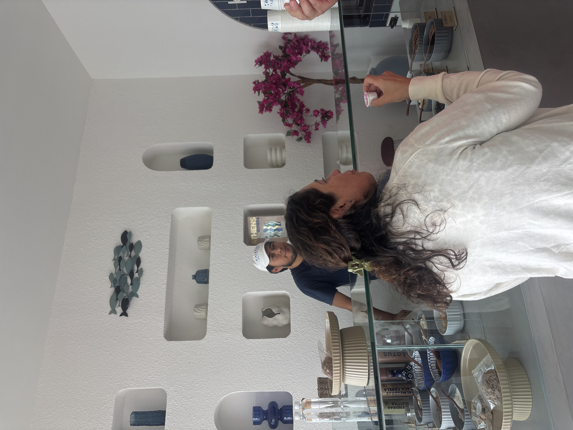 The image depicts the interior of a retail establishment, potentially a dessert shop or cafe, likely located in Puebla, Mexico, based on possible clues like the name "Athens" and the presence of Spanish language text. The interior design features a predominantly white color scheme with textured walls and decorative alcoves holding various objects, including dark blue pottery and a cluster of fish-shaped wall art. There's also a faux floral arrangement with pink flowers visible above a glass display case.
A woman with long, dark, curly hair is standing by the glass display case, seemingly reaching for a small container. Reflected in the glass is a man wearing a "Athens" hat, working behind the counter, potentially preparing or serving items. The display case contains various ingredients, potentially for desserts, in small bowls and containers. These include "Lotus", "Aca Canela", "Almendras Matcha". Some of the containers also have spoons in them.