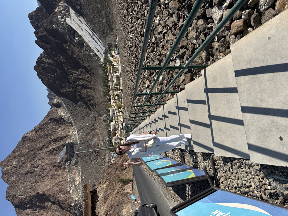 The image depicts an outdoor scene in Hatta, United Arab Emirates, characterized by rugged mountains and developed infrastructure, specifically in the vicinity of the Hatta Dam. The time of day is clear with strong overhead sunlight casting distinct shadows.
In the foreground, a female individual is positioned on a descending set of concrete steps, slightly left of the center. She is an adult, wearing a white crop top, white sweatpants, white sneakers, and a white vest draped over her shoulders. She also wears sunglasses and carries a light-colored crossbody bag. She faces slightly towards the camera, smiling, and holds her right hand up in a "peace" sign gesture.
To the immediate left of the steps, a paved two-lane road runs diagonally into the background. A dark-colored car is partially visible on the far left lane of this road. Several blue and white informational signs, supported by black frames and displaying text (partially visible as "KAYAK"), are positioned alongside the road. To the right of the steps, a dark green metal handrail parallels the descent, and the ground beyond it is covered with loose, irregular grey and brown rocks. Long, vertical shadows from the handrail posts and the individual are cast upon the concrete steps.
The midground reveals a valley floor containing a cluster of small, light-colored modular buildings interspersed with green trees and palm trees, indicating a developed recreational or accommodation area. Several streetlights equipped with solar panels are visible within this cluster. On the far right midground, a large, sloped concrete structure, likely a spillway or part of the Hatta Dam, extends down a mountain face. It features distinct rectangular patterns on its surface.
The background is dominated by towering, rugged brown and grey rocky mountains, rising steeply on both sides and receding into the distance. These mountains exhibit visible geological strata and jagged peaks. A winding road or path is visible traversing a distant mountain slope on the left. The sky above is clear and bright blue.