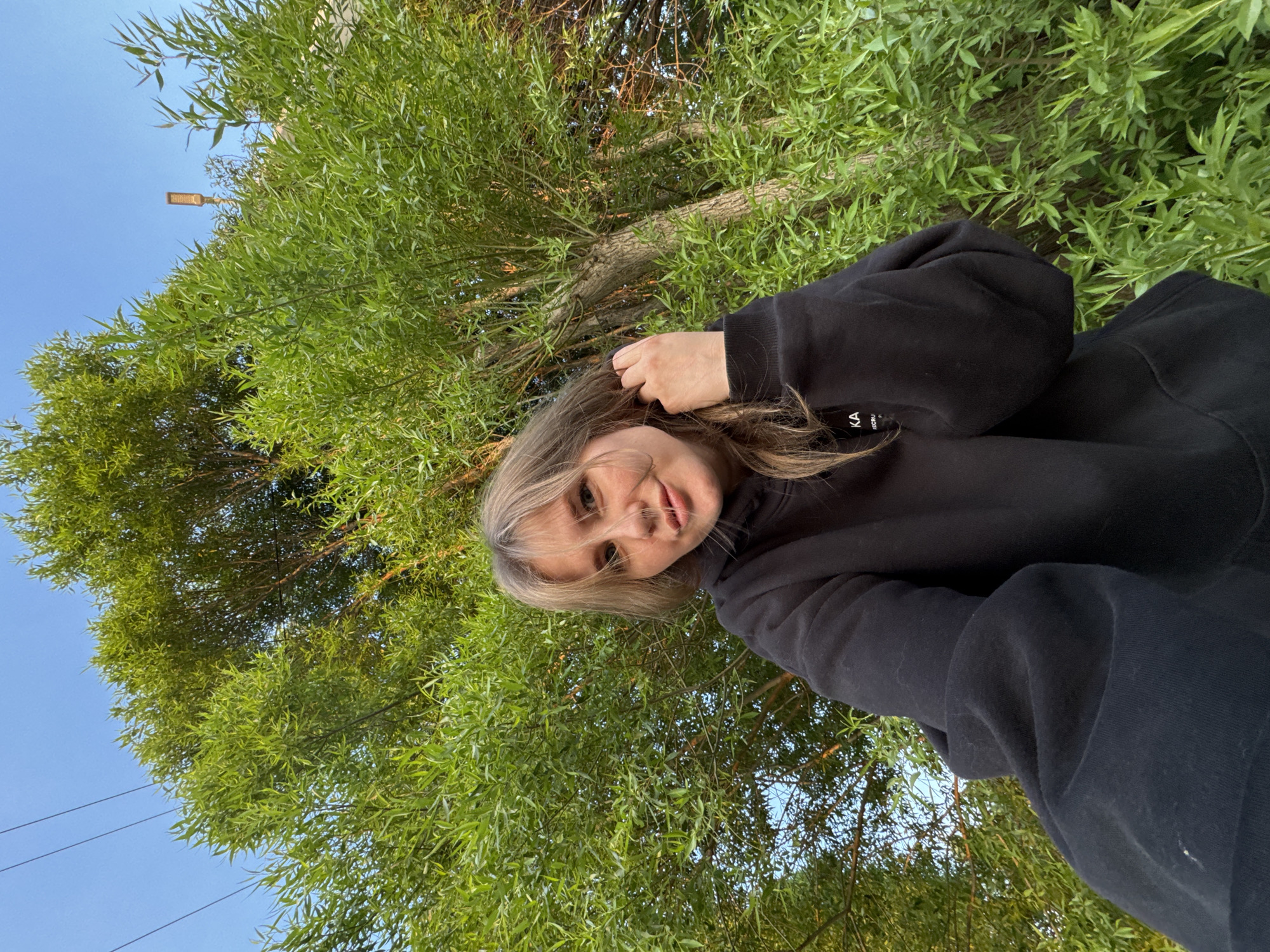 A woman in a black hooded sweatshirt is positioned in front of a backdrop of dense green foliage, likely from a tree. The scene includes a view of the sky, which is visible in the upper left corner. A utility pole and overhead wires can also be discerned against the sky. The location may be Горный, Russia, given the broader context.