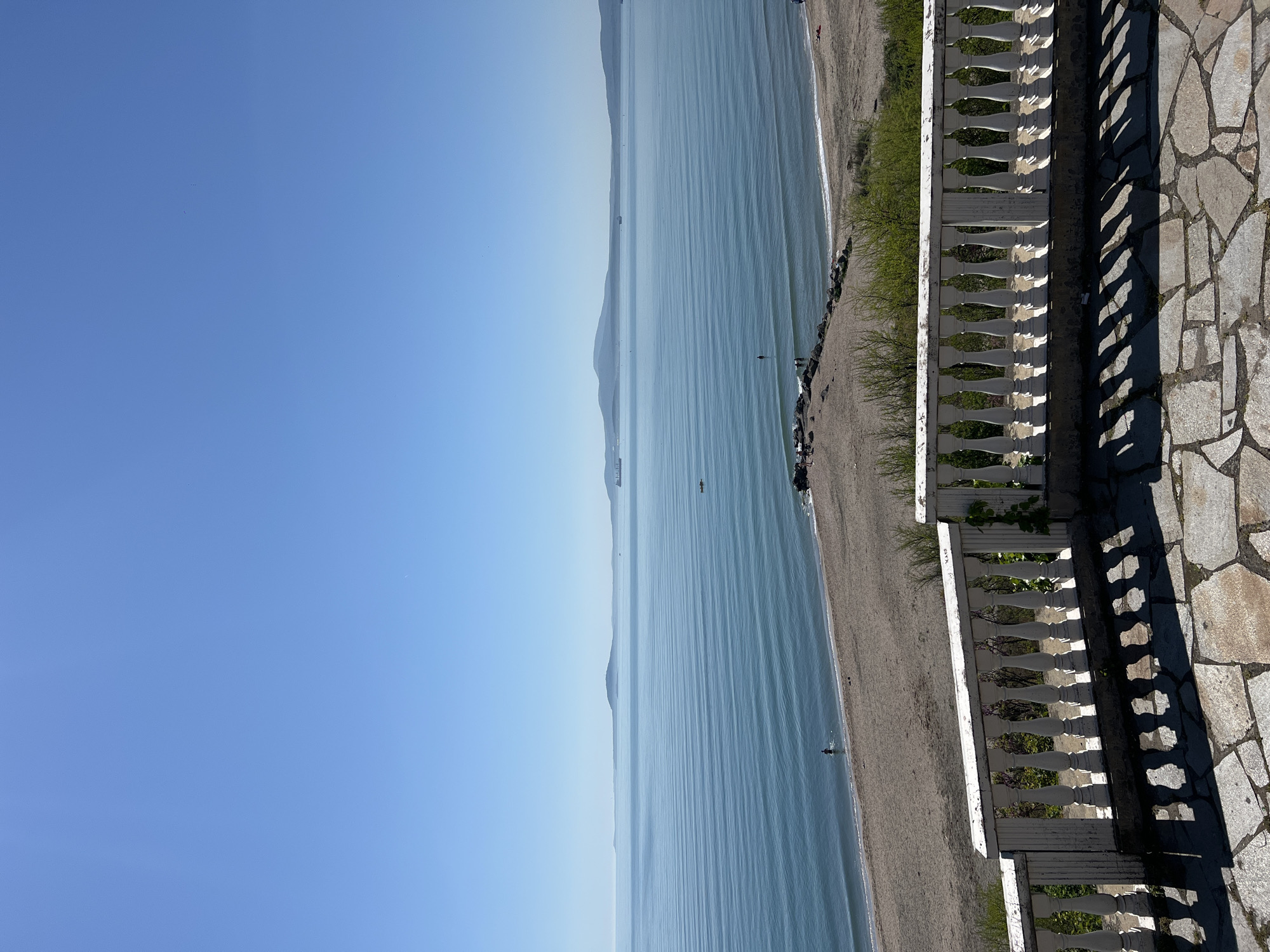 The image shows a view of the sea and coastline, possibly in Burgas, Bulgaria, as hinted by the user. The perspective is from slightly elevated, possibly from a balcony.
The sky is clear and blue, taking up about half the frame. The sea stretches into the distance, appearing calm with subtle lines on the water's surface. A long coastline runs along the right side of the frame. Along the coastline is a beach area, appearing sandy or pebbly. There are a few people visible on the beach.
A white, ornate balcony railing is present in the foreground, on the right side of the frame. To the right of the railing, there is a paved area, likely made of irregularly shaped stones. The angle of the sunlight casts distinct shadows from the railing onto the paved area.
