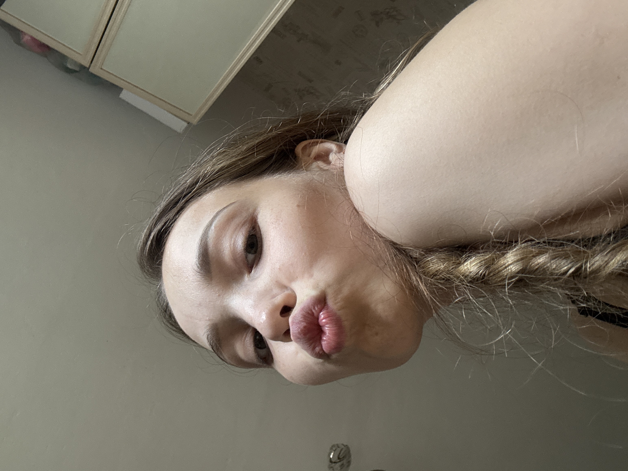 The image is a close-up, low-angle selfie of a young woman with blonde hair in a braid, puckering her lips. She is indoors, and the background appears to be a domestic setting with a wall and furniture. There is no identifiable information in the image that would suggest its location in Chaikovsky, Russia.