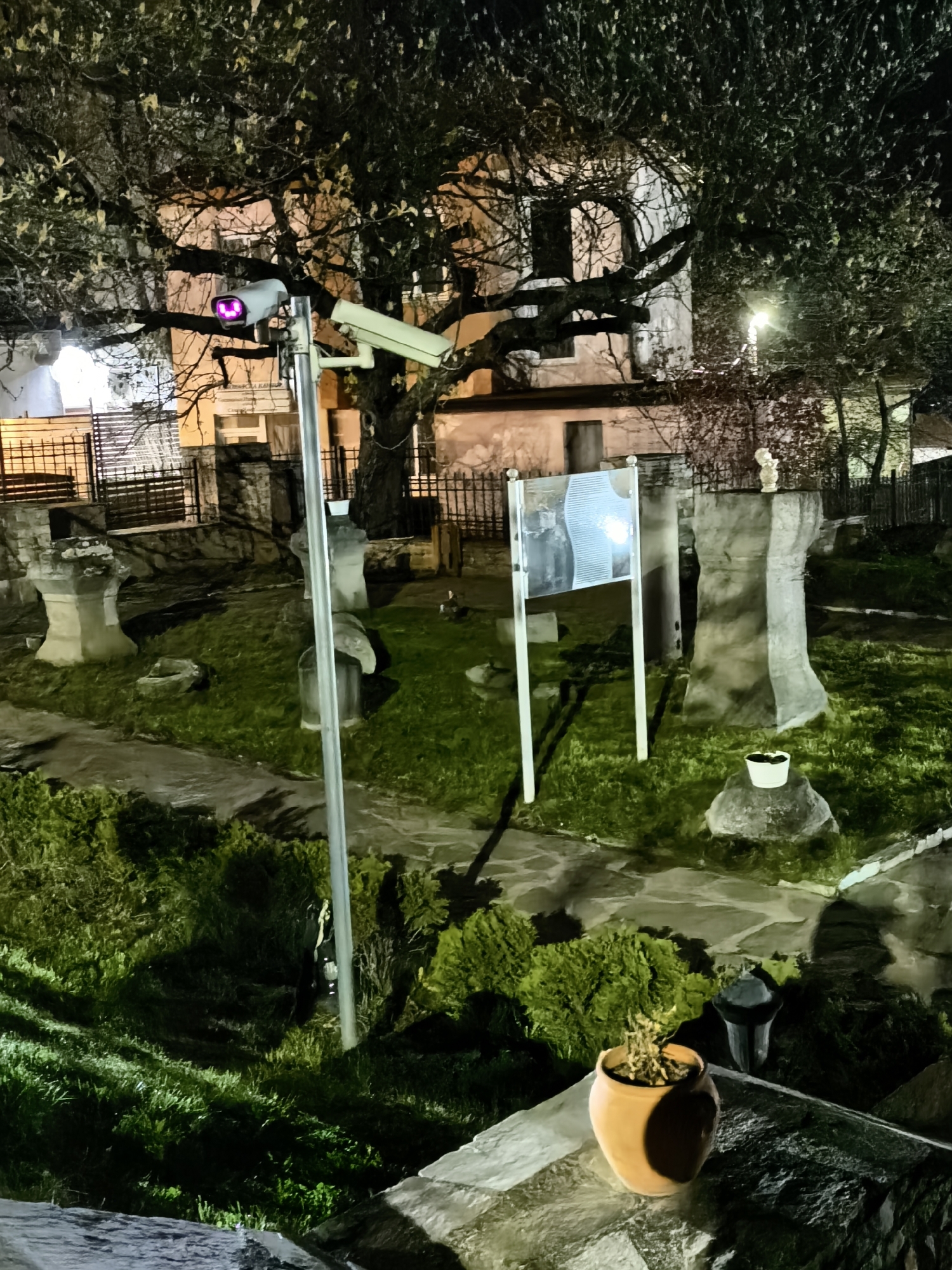 Captured at night, the photograph presents a location in the city of Unknown, Unknown. The scene is comprised of elements suggesting an outdoor area, possibly a garden or a historic site, illuminated by artificial lights.
A pivotal element is the presence of two security cameras, mounted on a pole, aimed to capture the surroundings. One camera is fixed, while the other is a cylindrical dome camera. They suggest monitoring and surveillance.
In the immediate foreground, there's a brown potted plant, sitting atop a stone surface. Further into the scene, there are stone structures, possibly monuments or architectural remnants, interspersed with patches of grass. A sign, seemingly displaying some form of information, is positioned centrally. The background is dominated by a large, bare tree with branches reaching towards the upper frame, with a building dimly visible behind.
The overall scene is quiet and static, with no evident human presence. The lighting is mixed, with some areas brighter than others, casting shadows that add depth to the image. The stone pathway and the greenery suggest a manicured and maintained outdoor space.