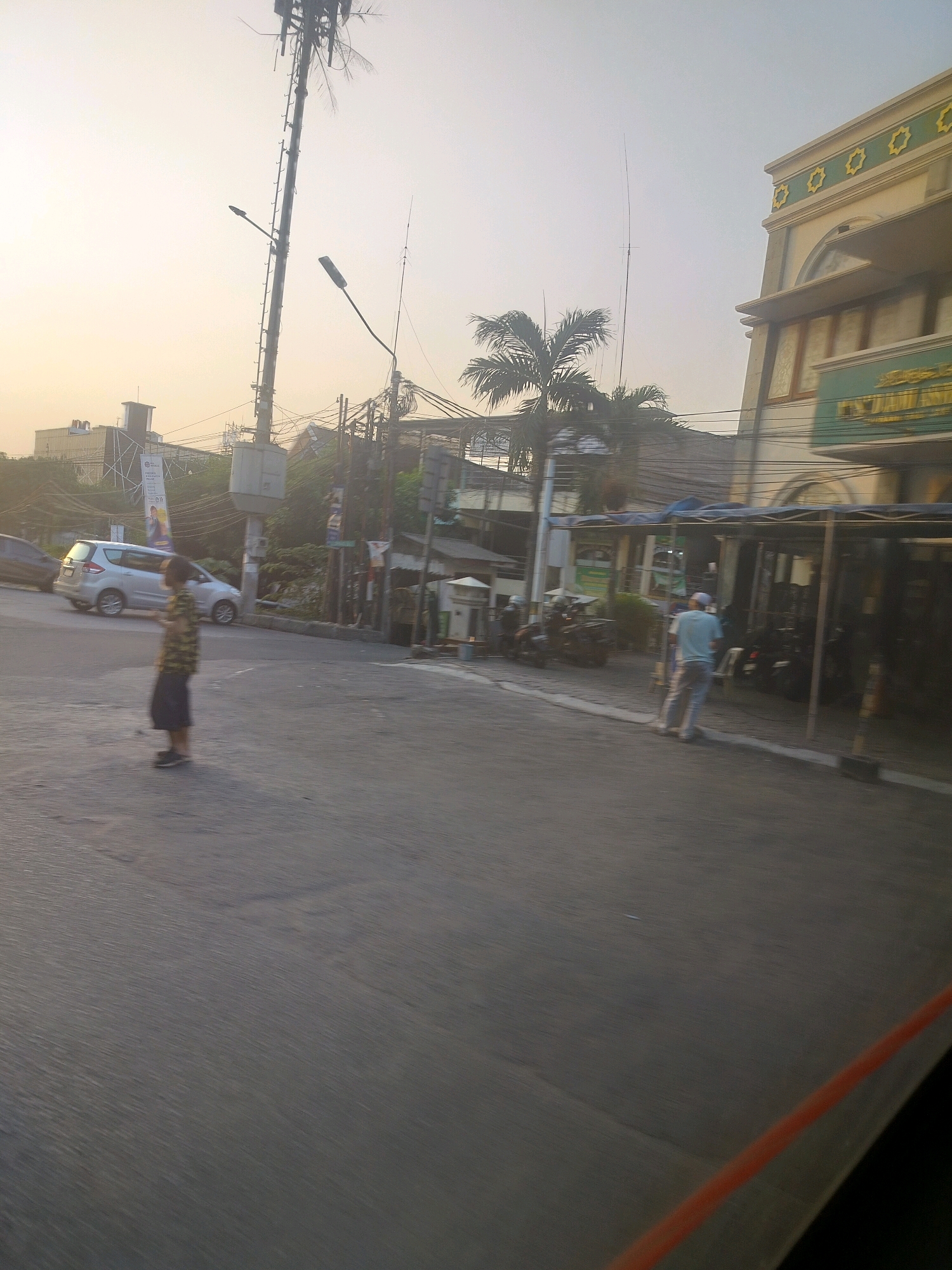 The image depicts a street scene in East Jakarta, Indonesia, likely during the late afternoon or early evening, given the soft, warm lighting and the long shadows. The foreground is dominated by asphalt road, showing signs of motion blur, suggesting the photo was taken from a moving vehicle.
On the left side of the street, a silver minivan is parked or moving slowly. In the middle ground, a young person, possibly a child, is walking across the road. They are wearing a patterned shirt and dark shorts. Further down the street, a man in a light blue t-shirt and grey pants is walking away from the camera towards a building. He appears to be wearing a cap.
Several vehicles, including motorcycles, are visible parked or moving near the buildings on the right. One building, possibly a mosque, is prominent on the right, featuring a cream-colored facade with decorative elements and arched windows. Another building on the left, with a simpler architectural style, has a banner with text and an image.
A tall communication tower stands in the background, with numerous wires and utility poles also present, characteristic of an urban environment. Palm trees are scattered throughout the scene, adding a tropical element to the urban landscape. The overall impression is a busy street intersection in a densely populated area of East Jakarta.