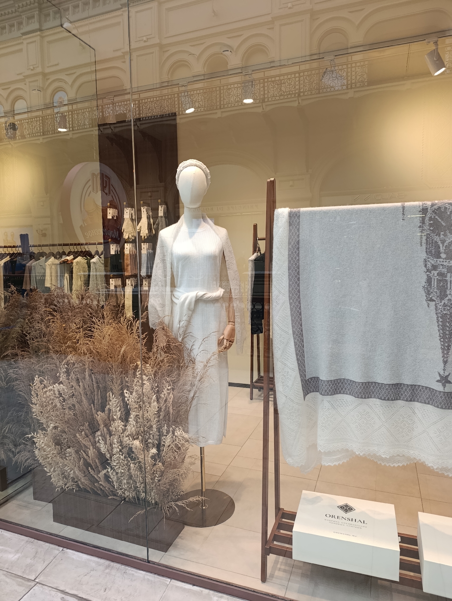 The image shows a storefront window display in Molokovo, Russia, featuring a mannequin dressed in white knit clothing, including a dress and a shawl. The mannequin is accessorized with a white headband and is positioned in front of a display of dried reeds. To the right, a large, light-colored textile with a grey border and intricate pattern, possibly a blanket or shawl, is hung on a wooden rack. Below the textile, on a shelf, is a white gift box with the brand name "ORENSHAL" printed in grey, along with Russian text indicating "FABRIKA ORENSBURGSKIH" and "PUSHOVYKH PLATKOV" (Orenburg Down Scarves Factory). In the background, a rack of clothing is visible, and the reflection of an ornate building interior, likely the GUM department store in Moscow, suggests the location might be within a larger shopping complex rather than a standalone store in Molokovo itself. The lighting is neutral, and the overall scene conveys a sense of retail display and product presentation.