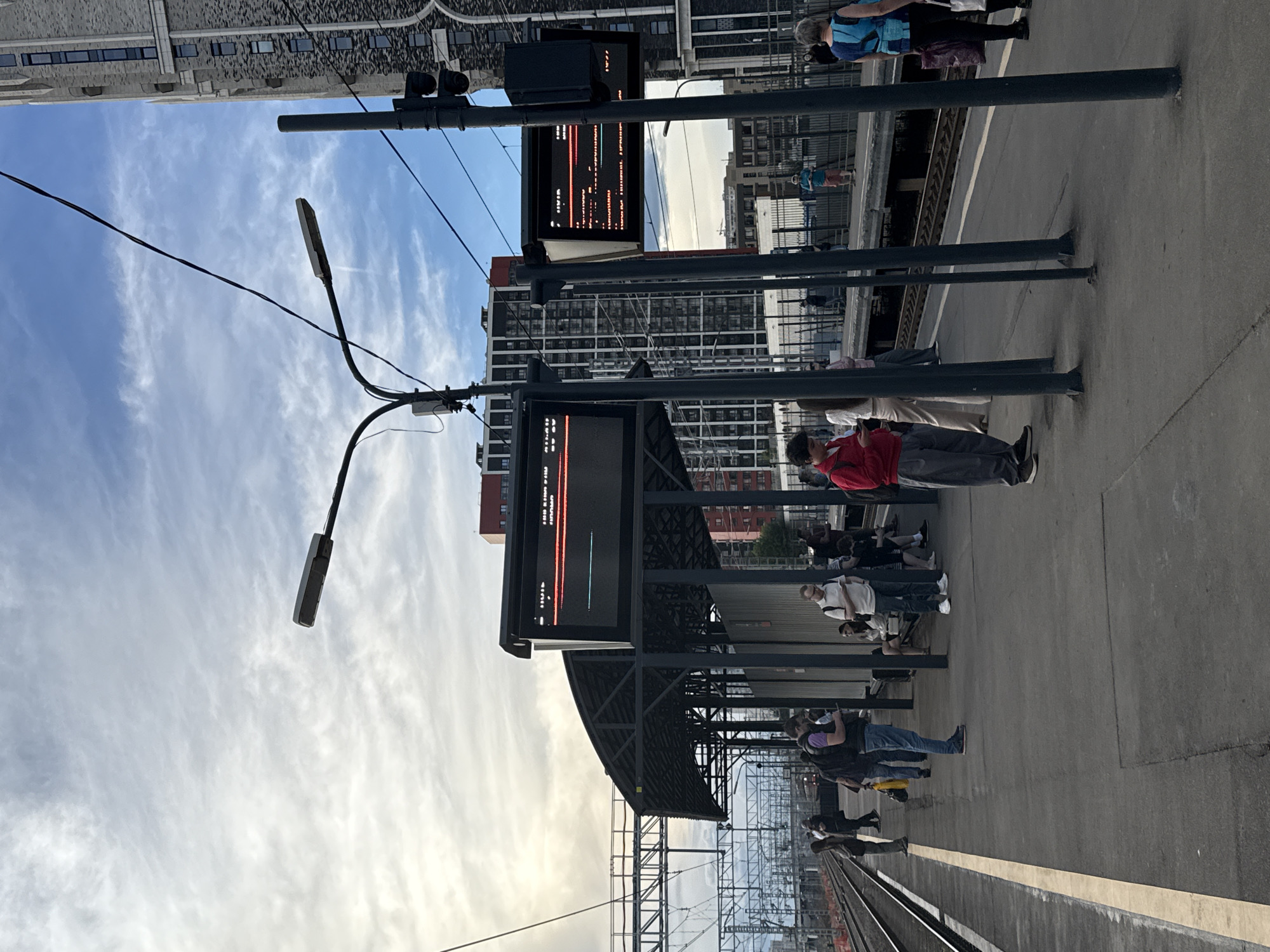 This media file captures a street scene in Moscow, Russia, likely at a train station. The image is oriented vertically, showing a platform with several people waiting. A prominent feature is a digital display board showing train information, with some text and graphics visible, including what appears to be arrival/departure times and destinations.
The weather appears to be cloudy, with a bright sky suggesting it might be daytime, possibly late afternoon or early evening, given the soft lighting. The scene is set on a concrete platform next to train tracks, with a covered waiting area supported by dark posts. In the background, there are modern buildings, including a tall, glass-fronted structure and other multi-story buildings.
The people are dressed casually and appear to be waiting patiently for their trains. Some are standing, while others are seated on benches under the shelter. Their expressions are neutral, indicating a common commuter or traveler atmosphere. The overall mood is calm and functional, typical of a public transportation hub. The presence of electrical wires and streetlights further confirms the urban setting.