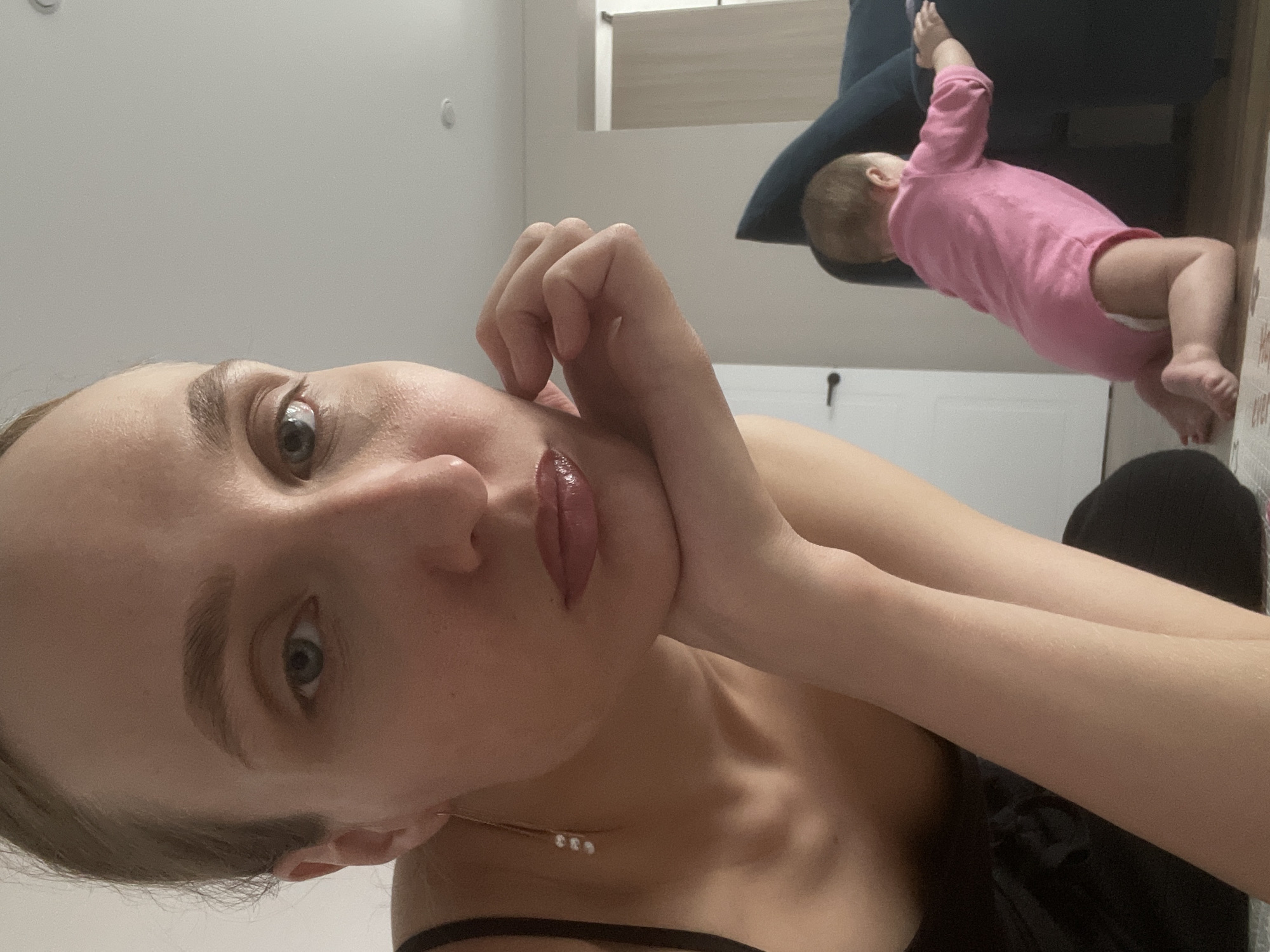 An image, presented vertically, captures a candid indoor scene featuring a woman in the foreground and a baby in the background. The woman, with light hair and eyes, lies with her head resting on her hand, gazing directly at the camera. She wears a black strap top and a delicate necklace featuring three small, shiny elements. Her lips are adorned with a glossy, dark pink color.
Behind her, a baby, dressed in a pink long-sleeved bodysuit, is visible on its stomach, with one arm extended upwards towards a dark blue cushion or piece of furniture. The baby's head faces away from the camera. The background consists of light-colored walls or flooring, a partially visible white doorframe, and a small dark keyhole or switch on the wall. Underneath the baby's feet, there is a textured surface with some faint, illegible light-colored text or pattern.
The scene suggests a quiet, domestic moment, with soft, ambient indoor lighting. The woman's expression appears calm or thoughtful. There are no specific visual cues within the image to indicate its location as Sepych, Russia.