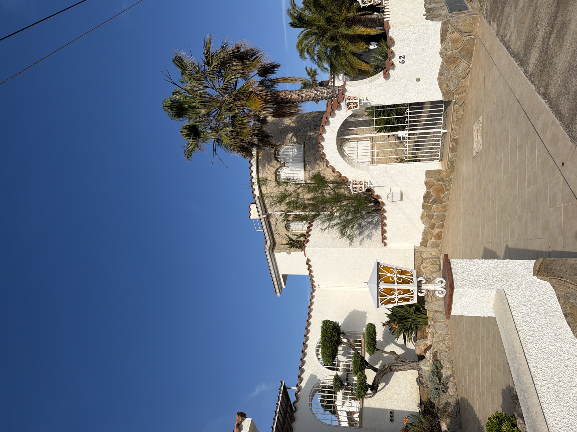 This media file captures the exterior of a Mediterranean-style building under a clear, vibrant blue sky, indicative of Mont-roig del Camp, Spain. The scene is brightly lit by the sun, with distinct shadows suggesting it is either morning or late afternoon.
The building features white, textured stucco walls, complemented by decorative terracotta roof tiles along its eaves and curved architectural details. Portions of the facade are adorned with natural, irregularly shaped light brown and grey stone cladding. Several arched windows are visible, each fitted with ornate white metal grilles.
Prominently featured are several palm trees, with one large, mature specimen dominating the mid-ground, its trunk and fronds clearly visible against the sky. Various other plants enhance the landscaping, including smaller bushes, a carefully sculpted, bonsai-like shrub, and bird of paradise plants.
An ornate white lamppost with amber-colored glass panels stands near the front. White metal gates provide access to the property, and a white mailbox is mounted on the wall. The ground in front of the house is paved with light brown square tiles, leading to a darker asphalt area, likely a road or driveway, on the right. The number "62" is clearly visible on the white wall adjacent to an entrance gate. Overhead, faint power lines stretch across the upper left portion of the deep blue sky. The overall impression is that of a peaceful, well-maintained residence in a sunny, pleasant environment.