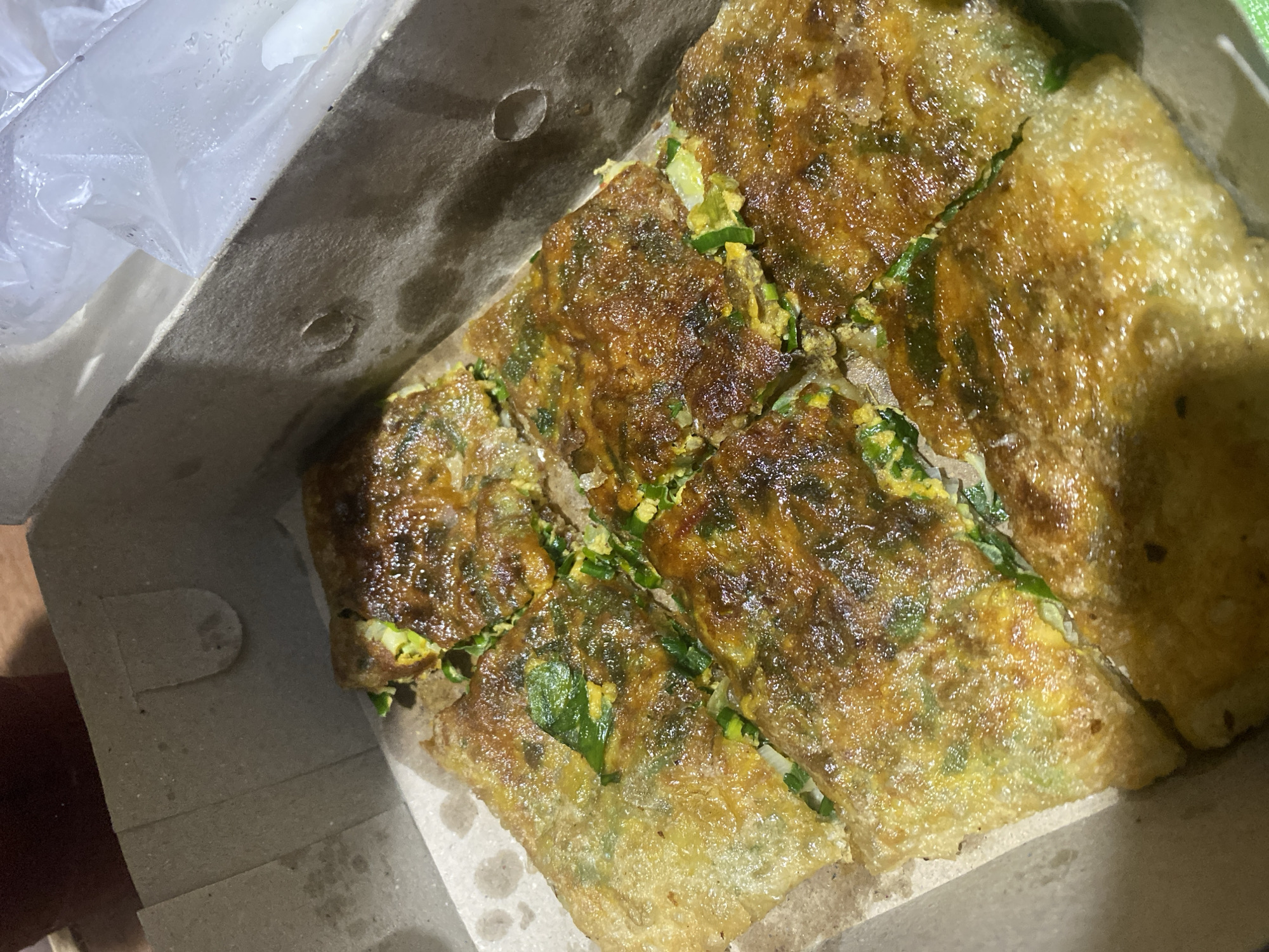 A close-up, overhead shot shows six pieces of savory martabak, a fried stuffed pancake, arranged in a cardboard takeout box. The martabak pieces are golden brown and appear to be filled with a mixture of eggs and green onions. The box is open, and a blurred white plastic bag is visible in the upper right corner. The background is out of focus, but suggests a wooden surface. The scene is indoors, likely in a home or food stall in Ta', Indonesia, given the context.
