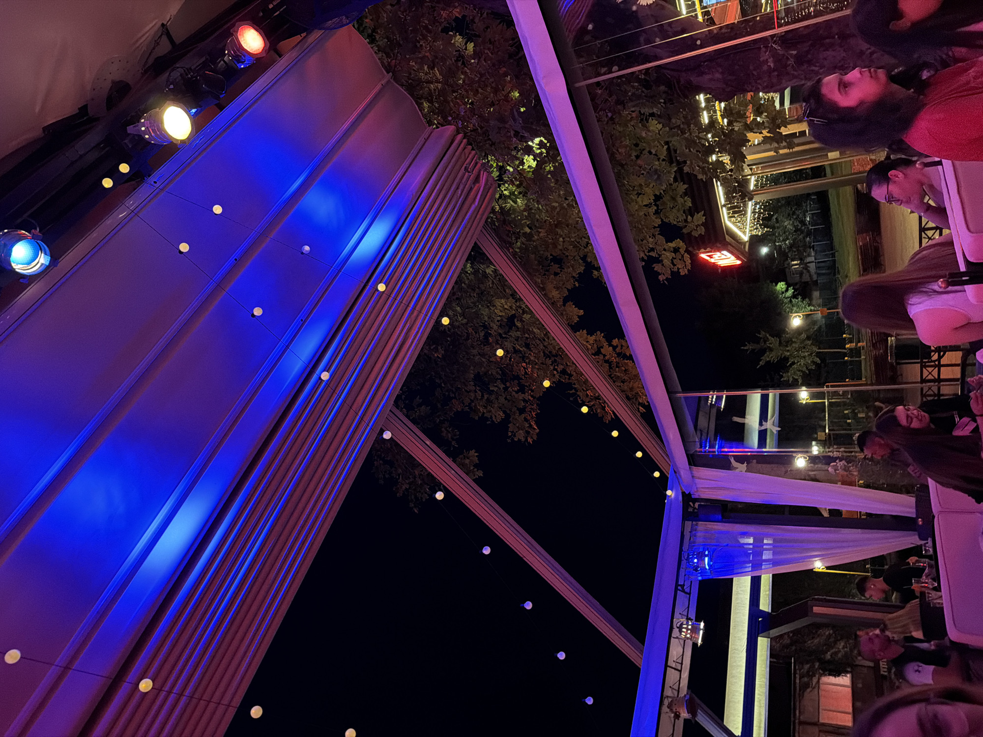 The image depicts an outdoor seating area at night, likely a restaurant or bar, with a retractable roof structure illuminated by blue and pink lights. Small, round yellow lights are strung across the ceiling, and larger spotlights are also visible. Trees with foliage are seen in the background, with a neon "HOTEL" sign also illuminated. Several people are seated at tables, some of whom are engaged in conversation. The scene suggests a lively evening atmosphere. The location is identified as Haskovo, Bulgaria.