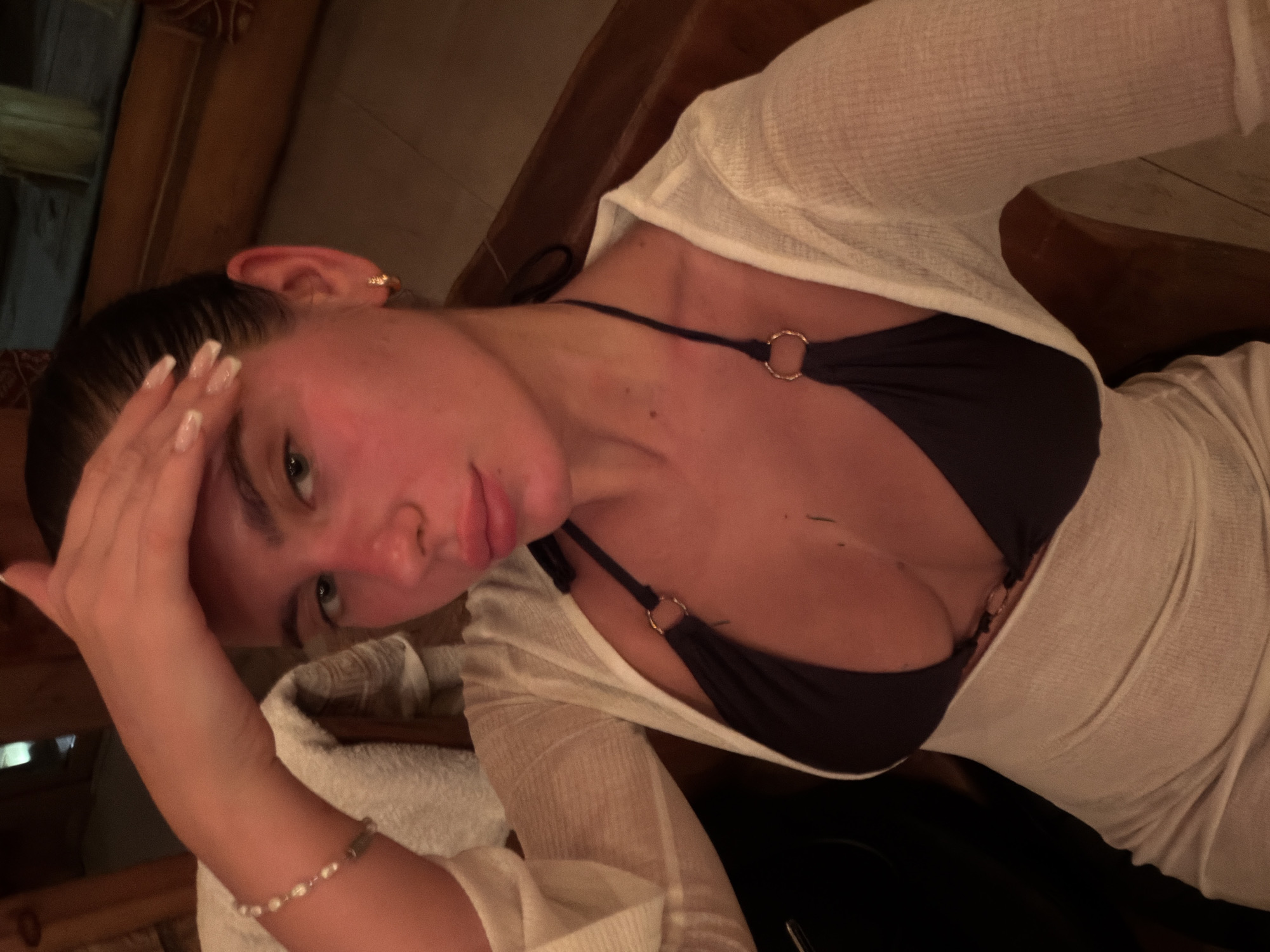 The image captures a close-up, tilted portrait of a young woman with fair skin and dark hair pulled back. Her face appears slightly flushed, and she looks directly at the camera with a neutral to slightly pensive expression, her lips parted. She rests her right hand on her forehead, revealing long, manicured fingernails with French tips.
The woman is dressed in a dark-colored bikini top or bralette, featuring a prominent gold O-ring detail on the strap. Over this, she wears a sheer, white or off-white, long-sleeved cover-up or top. A delicate beaded bracelet is visible on her left wrist, and a small gold earring adorns her right ear.
The setting, located in Drokino, Russia, appears to be an indoor space characterized by warm, dim lighting, suggesting an evening or nighttime scene. The background consists of earthy, wooden tones, possibly indicating wood paneling or furniture, hinting at a cozy room, spa, or sauna environment. A white towel is visible near her left arm. The overall atmosphere is intimate and relaxed, with the soft lighting highlighting her features. No visible text is present in the image.