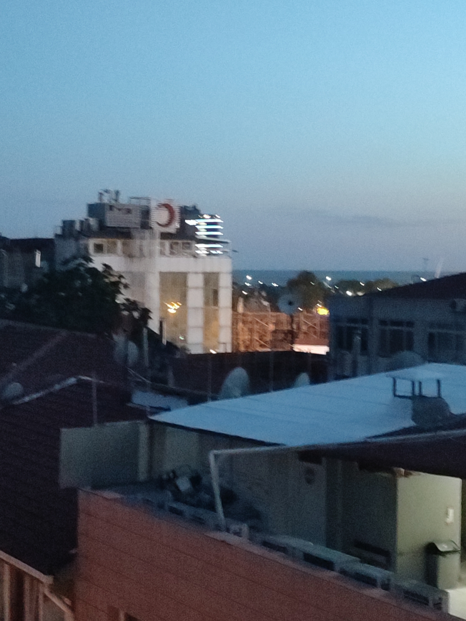 This image depicts a dusk or twilight scene in Kadıköy, Turkey, showcasing a mix of urban architecture against a muted sky. In the mid-ground, a multi-story building with a prominent red crescent logo on its upper section is visible. The building has several windows and a large, brightly lit vertical section. To the right of this building, another structure with a darker facade and rectangular windows is present. In the foreground, rooftops of residential or commercial buildings are visible, one with a red-tiled roof and another with a lighter-colored corrugated metal roof. The background suggests a body of water, likely the Sea of Marmara, with distant lights dotting the horizon. The overall atmosphere is calm, with the fading light creating a serene urban landscape.