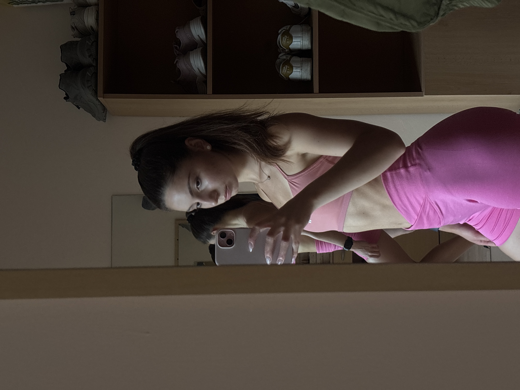 A young woman with dark hair pulled into a high ponytail stands in front of a full-length mirror, taking a selfie with her phone. She is wearing a pink sports bra and matching pink shorts. Her reflection is visible in the mirror, showing her from the side. The background includes a wooden shelf unit filled with shoes. The room appears to be a bedroom or dressing area. The overall lighting is dim. There is no indication of the city of Varna, Bulgaria.