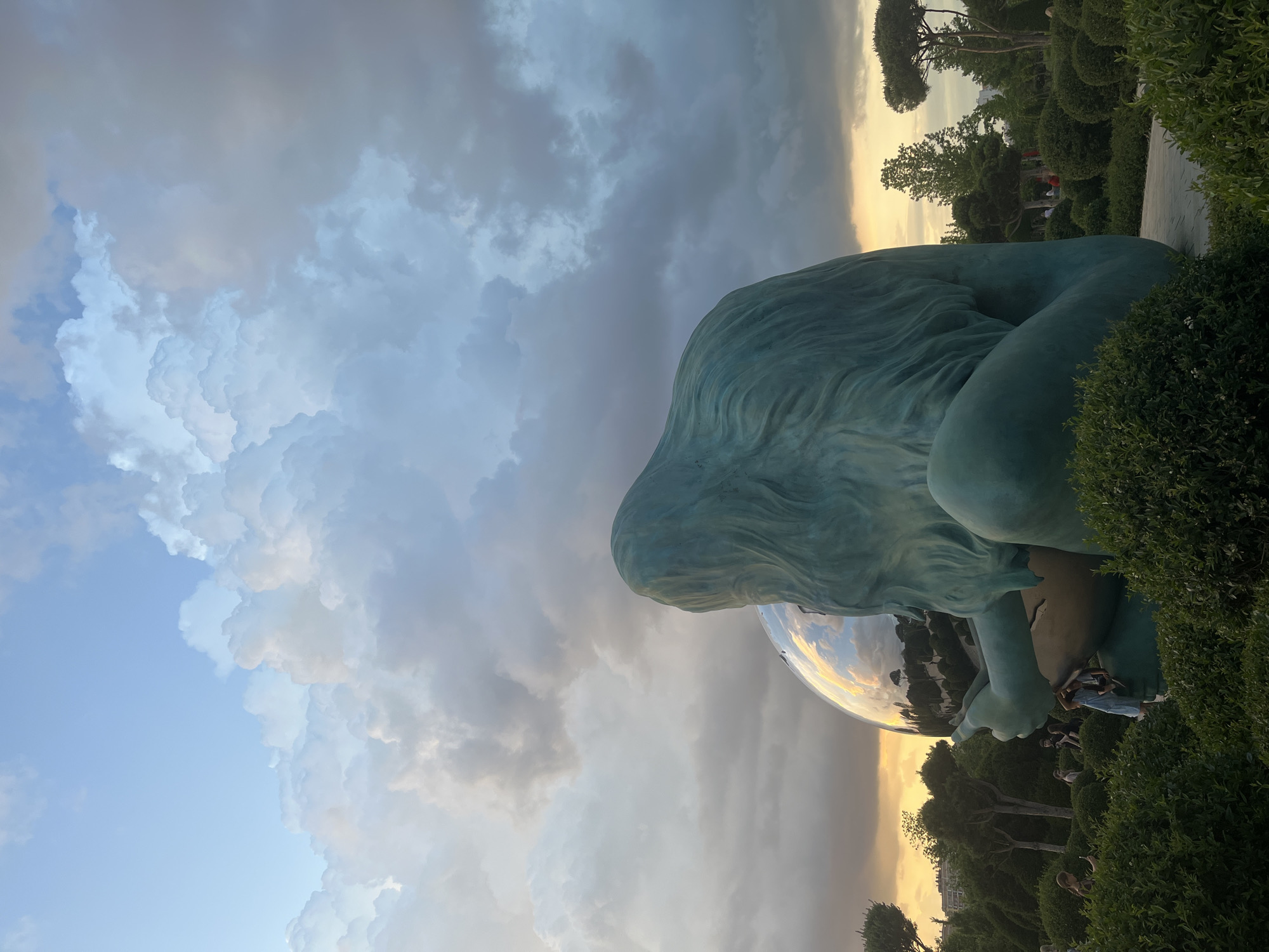 The image depicts a large, teal-colored sculpture of a seated figure, seen from behind, with long, flowing hair. The figure is cradling a large, highly reflective, spherical object in its hands. The sculpture is situated outdoors in a park-like setting with manicured bushes and trees. In the background, there are buildings and more trees. The sky above is filled with dramatic, billowing clouds, with a sunset or sunrise visible near the horizon, casting a warm glow. Several small figures, appearing to be people, are visible in the middle ground, some interacting with the sculpture and others walking through the park. The scene appears to be illuminated by natural light. This sculpture is located in the city of Krasnodar, Russia.