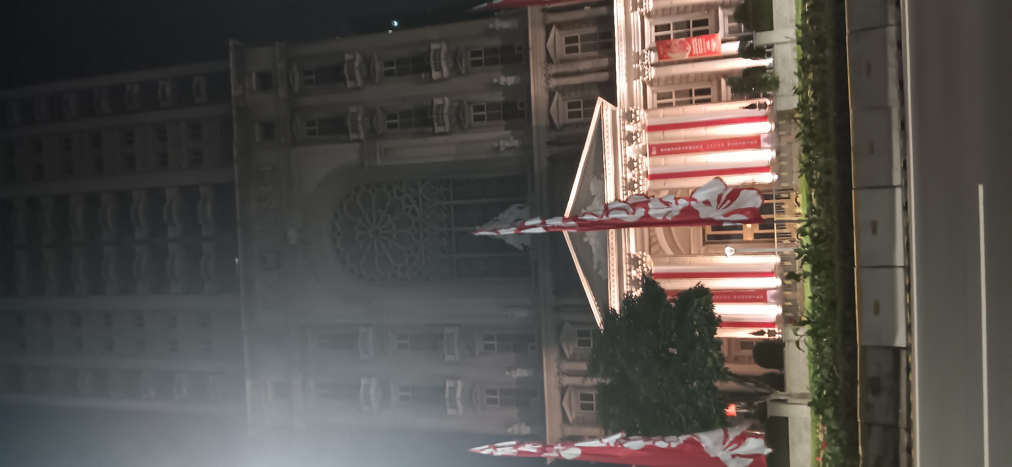 This is a nighttime exterior, ground-level shot of the facade of a grand building, likely a hotel or a historic monument, situated in Central Jakarta, Indonesia. The building features classical architectural elements, including columns, intricate window designs (a large rose window is visible), and balconies. The facade is illuminated, creating a dramatic effect. Red and white banners, possibly national flags or decorative elements, hang prominently in front of the building, some draped vertically and others as tall flags on poles. A tree is situated to the left of the entrance, and manicured greenery lines the front of the property. In the foreground, a road with visible lane markings and concrete barriers separates the viewer from the building. The scene is slightly hazy, possibly due to mist or light diffusion. No people are clearly visible or engaged in any discernible activity.