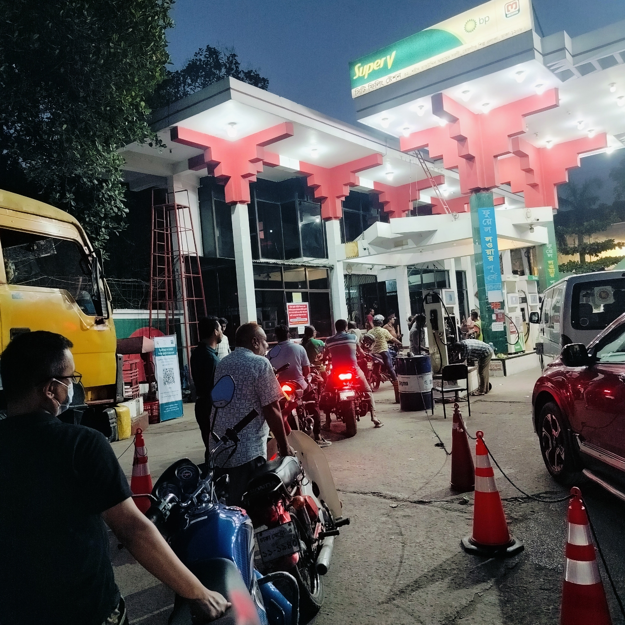 The image shows a busy BP gas station with the "Super V" logo at night in Dhaka, Bangladesh. Several people on motorcycles and in cars are waiting in line for gas. A yellow truck is on the left side of the image. The license plate number of one of the motorcycles is "ঢাকা মেট্রো-এ ১১-১৯৪০". Orange traffic cones mark the waiting area. The gas station’s building has a red and white color scheme. A ladder is standing against the wall of the gas station.
