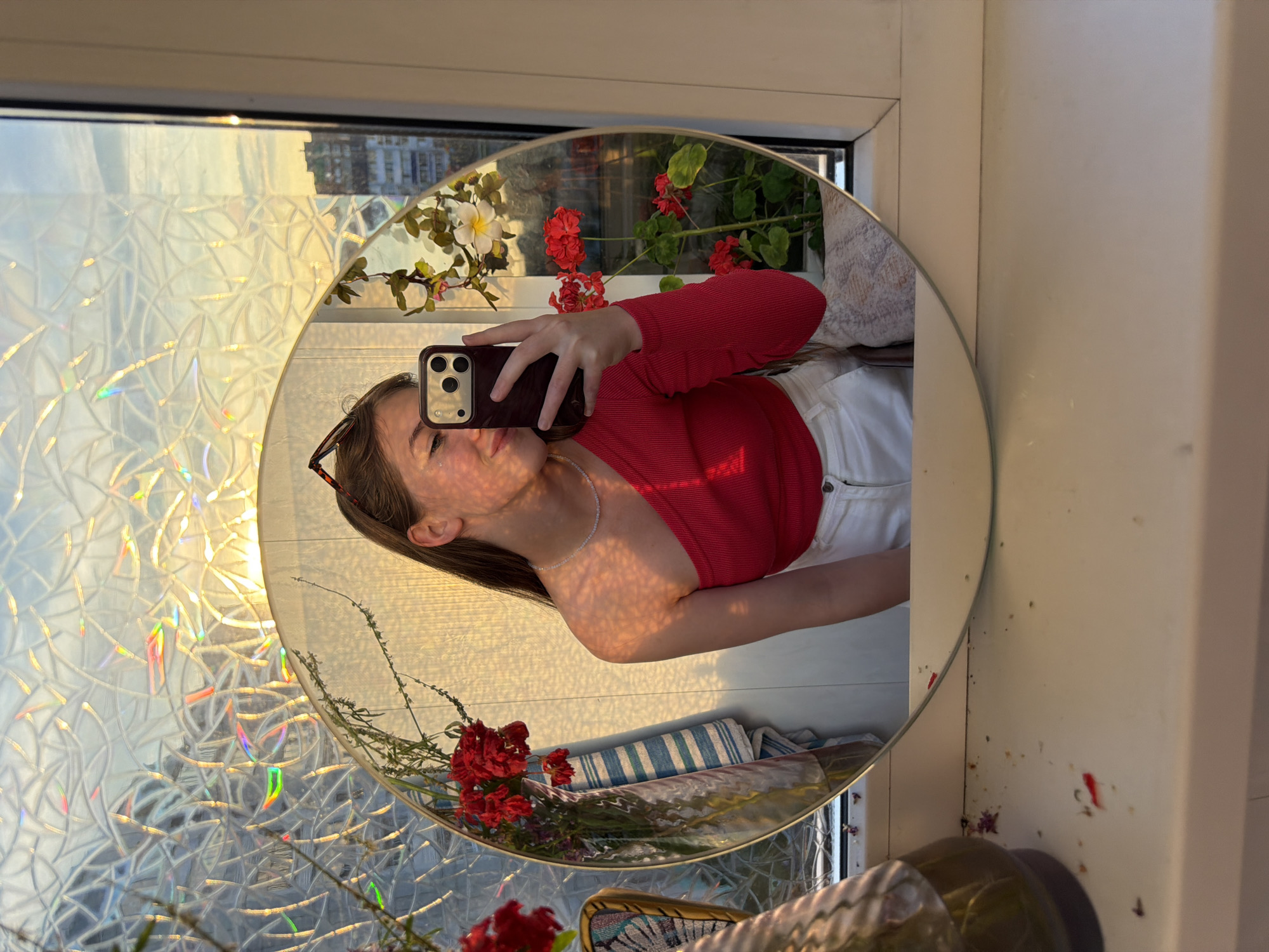 A young woman with long brown hair, wearing a red one-shoulder top and white pants, is taking a selfie in a circular mirror. She is holding a dark-colored smartphone in her right hand. She is smiling and her left cheek is lightly touching her hand. She is wearing a delicate pearl necklace. The mirror reflects her and the surrounding environment. Behind her, a patterned window with sunlight filtering through casts iridescent colors. There are red flowers in a vase to the left and a potted plant with red and white flowers to the right, both reflected in the mirror. The scene appears to be indoors, possibly on a balcony or near a window, with a view of buildings in the distance. The overall lighting suggests it is either sunrise or sunset. The windowsill beneath the mirror is cluttered with small debris, including what appear to be dried flower petals and small bits of red material. The image was captured in Chaikovsky, Russia, specifically in an apartment.