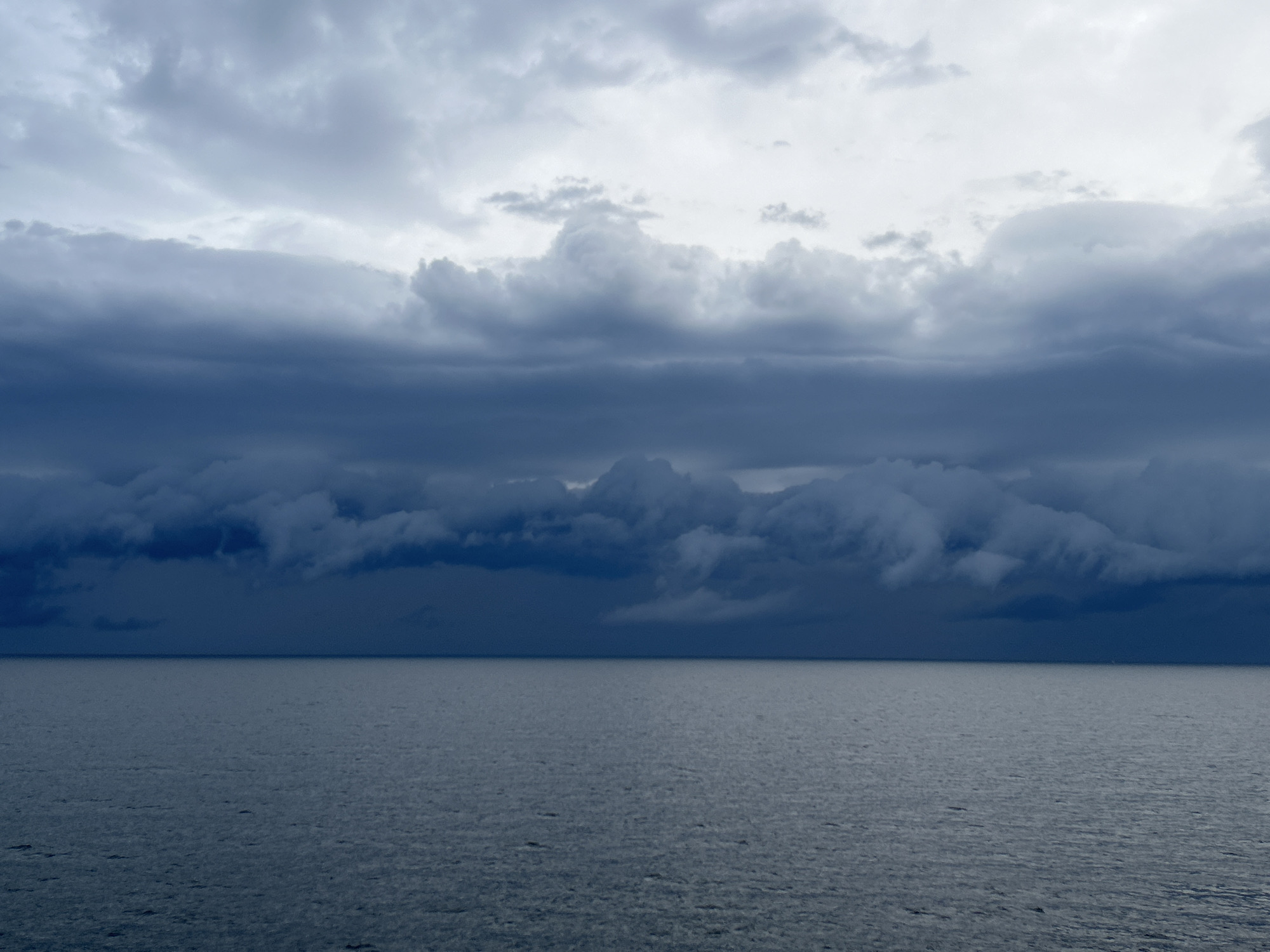 The image captures a dramatic seascape under a heavily clouded sky. The scene is divided horizontally between the vast expanse of water in the lower half and the layered clouds occupying the upper half.
The sky is dominated by shades of grey and dark blue. The uppermost layer features lighter, scattered grey clouds, suggesting some light breaking through or diffuse light. Below this, a thick, horizontal band of dense, dark grey and blue-grey clouds stretches across the middle of the sky. These clouds appear heavy and turbulent, particularly at the horizon line where they become almost black, indicative of an approaching storm or very overcast conditions. There's a subtle brighter area in the upper-right sky, hinting at a light source behind the clouds.
The body of water below is a uniform dark blue-grey, mirroring the somber tones of the sky. The water's surface is relatively calm, with small, subtle ripples visible, suggesting a gentle movement but no significant waves. The horizon line is perfectly flat and clear, creating a stark separation between the heavy sky and the serene, vast ocean.
There are no visible people, objects, or landmasses in the image. The time of day appears to be overcast daytime, possibly late afternoon or early morning given the diffuse light, with the overall weather suggesting impending rain or a storm. The scene evokes a sense of immense scale, tranquility, and potentially a foreboding atmosphere due to the dark clouds. There is no visible text.