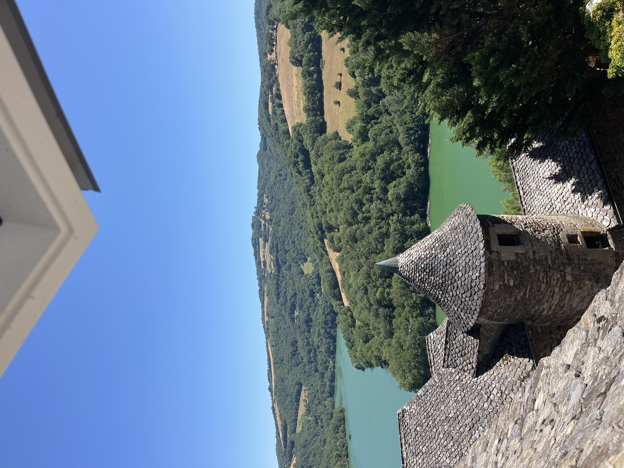 This media file captures a scenic view of a lake and surrounding landscape, with a castle structure in the foreground. The scene appears to be set on a clear, sunny day, likely in the late morning or early afternoon, given the bright light and blue sky.
The main subjects include a tranquil, green lake, a densely forested hillside rising from the lake's edge, and a section of the lake's opposite shore with cultivated fields and scattered trees. In the foreground, a portion of a stone castle is visible, featuring a distinctive conical roof covered in grey, scale-like tiles. The castle walls are constructed from rough-hewn stone. Two windows are visible in the stone tower.
There is no discernible activity or event taking place. The focus is on the serene natural beauty of the landscape and the imposing presence of the historical castle. The location cues suggest a European setting, possibly a mountainous or hilly region. The overall mood conveyed is one of peace and historical grandeur. No text is visible in the image. The orientation of the image is such that the landscape is viewed from a high vantage point, looking down towards the lake and across the valley. The white structure on the left edge of the frame is likely part of the same castle complex, with a distinctive angled roofline.