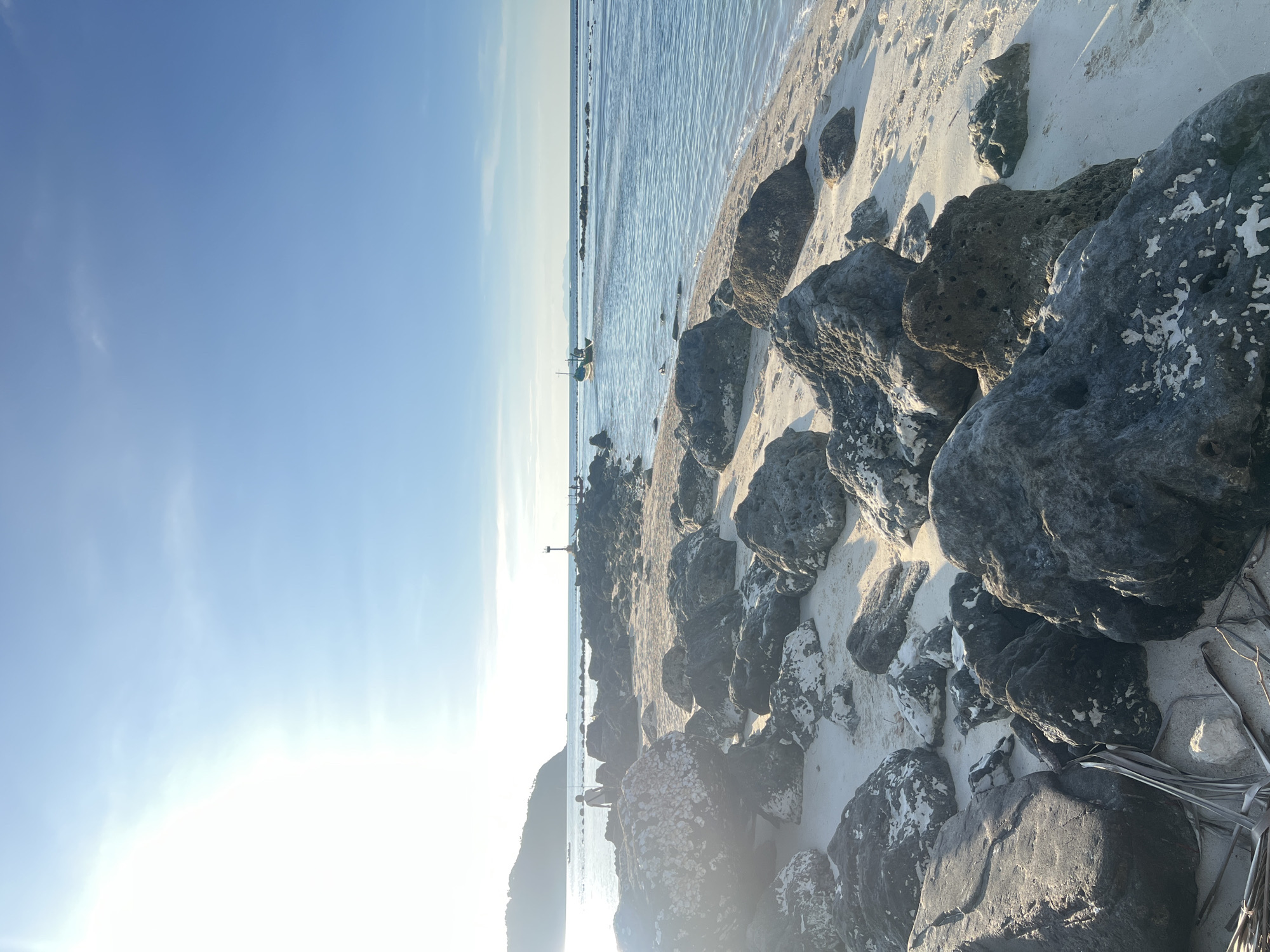 The image captures a serene beach scene on Koh Phangan, Thailand, likely in the vicinity of Tambon Ko Phangan. The foreground is dominated by large, dark grey rocks scattered across a sandy beach, with some white mineral deposits visible on their surfaces. In the middle ground, a rocky jetty or breakwater extends into the clear blue ocean, with a small beacon visible on it. A person is standing near the beginning of this jetty, facing the water. Further out in the calm sea, a small, traditional Thai fishing boat with a single occupant is heading towards the horizon. The sun is low in the sky, casting a bright glare on the left side of the image and creating a warm, golden light that reflects off the water and illuminates the landscape. The sky is a clear, pale blue with a few wispy clouds. In the background, a lush, green, mountainous island rises from the sea. The overall atmosphere is one of tranquility and natural beauty, characteristic of a tropical beach setting.